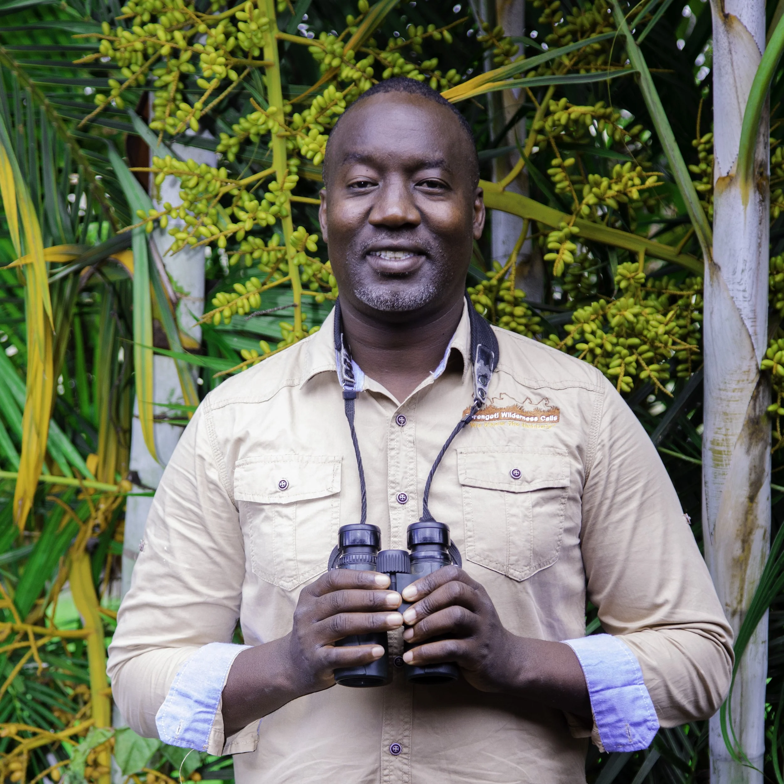 A man standing outdoors in front of green foliage, holding binoculars. He's wearing a beige long-sleeved shirt with a logo on the chest and has a black lanyard around his neck.