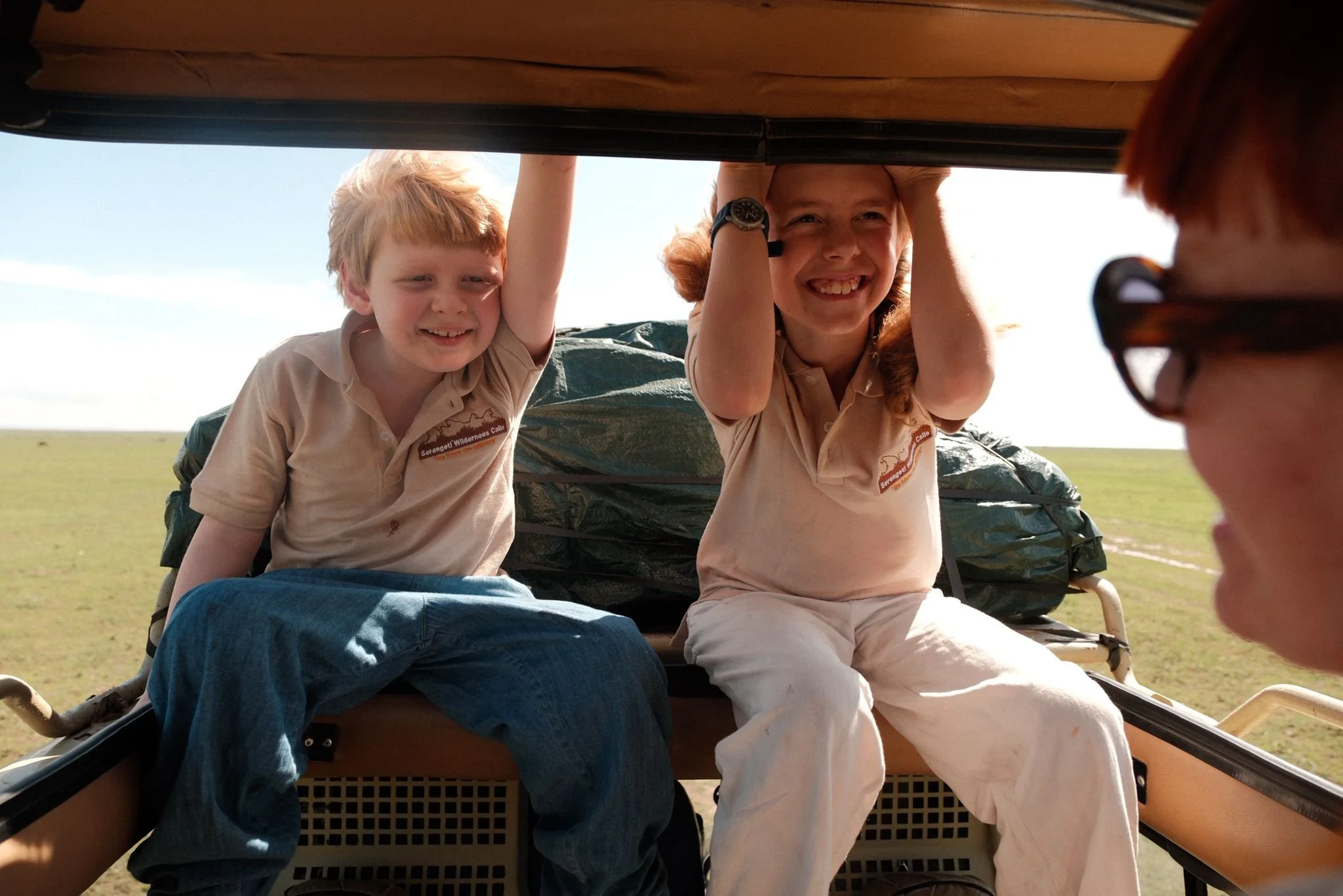 Two children and a guide look out from the back of a safari vehicle in an open grassland. The children are smiling and looking over the edge of the vehicle, enjoying a wildlife safari experience.