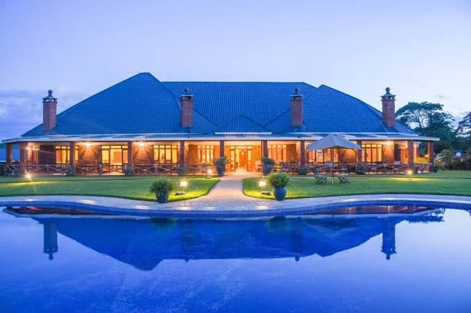 A large house with a blue roof and illuminated windows, surrounded by a well-maintained lawn, potted plants, and outdoor furniture. There is a swimming pool in the foreground reflecting the house.