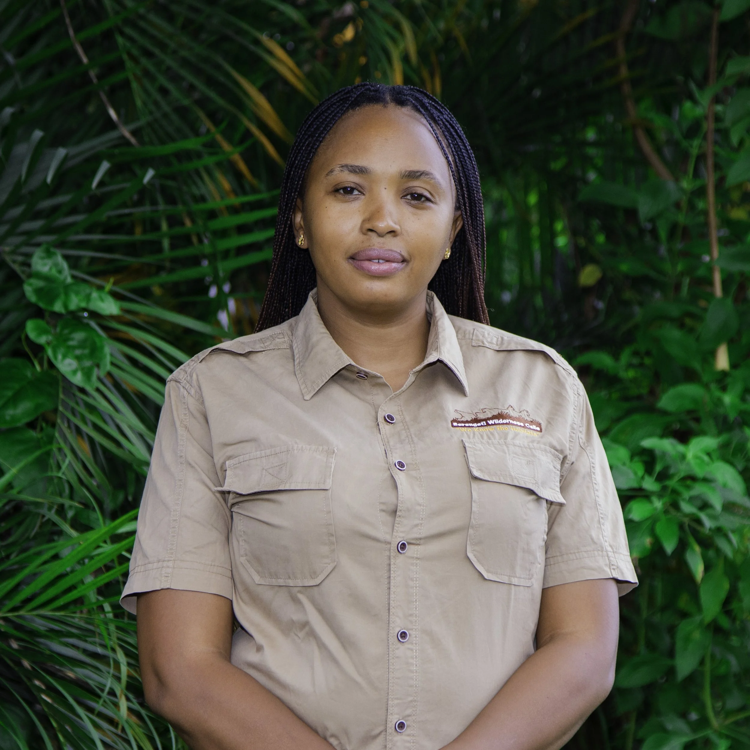 A woman in a khaki uniform standing outdoors against a background of green tropical plants.