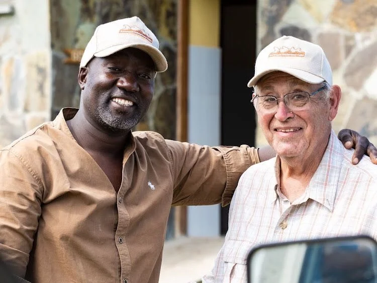 Two smiling men wearing baseball caps, standing close together with their arms around each other, outdoors in front of a stone building.