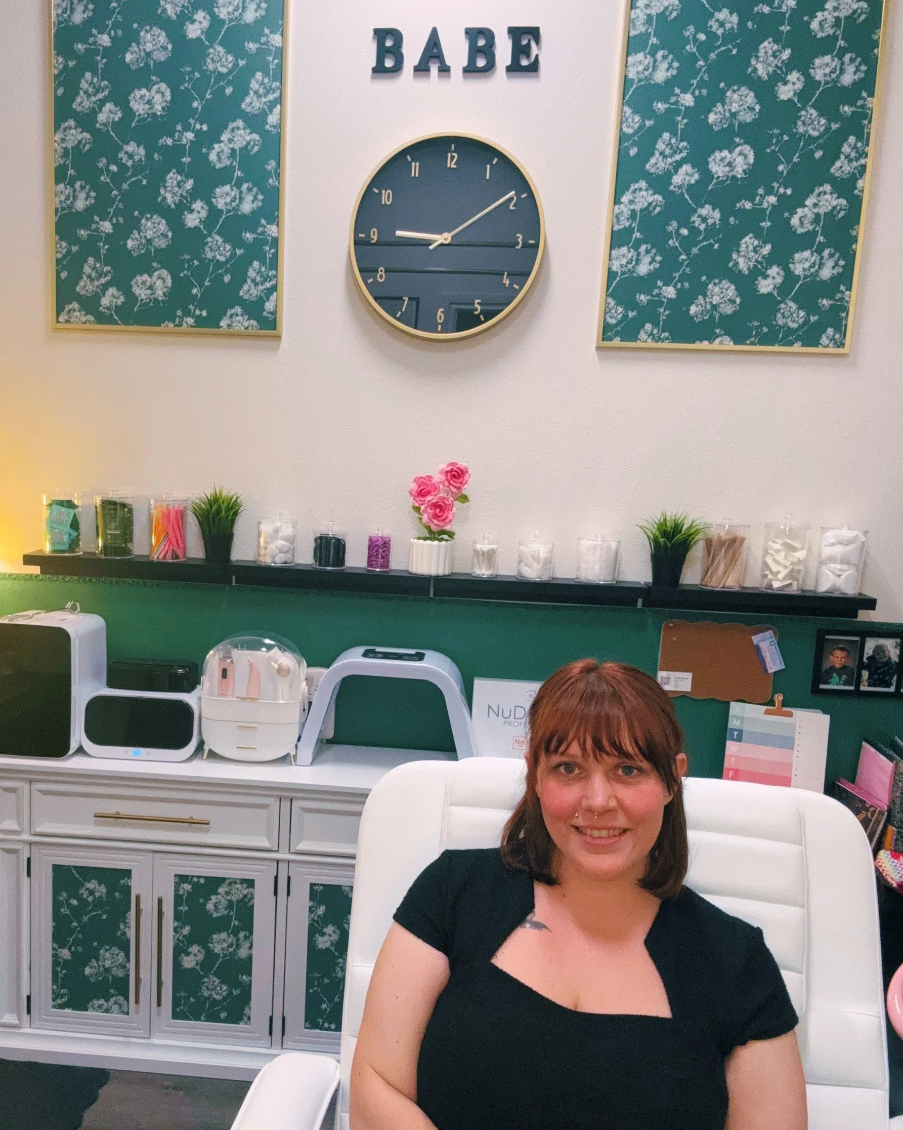 A woman sitting in a white chair, smiling, in a room with green and white floral wallpaper on the cabinet behind her, a clock on the wall above the cabinet, and shelves with decorative items, including pink flowers and various supplies.