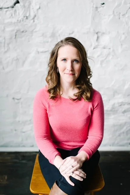 Woman with long wavy hair wearing a pink sweater sitting on a wooden chair in front of a white brick wall.