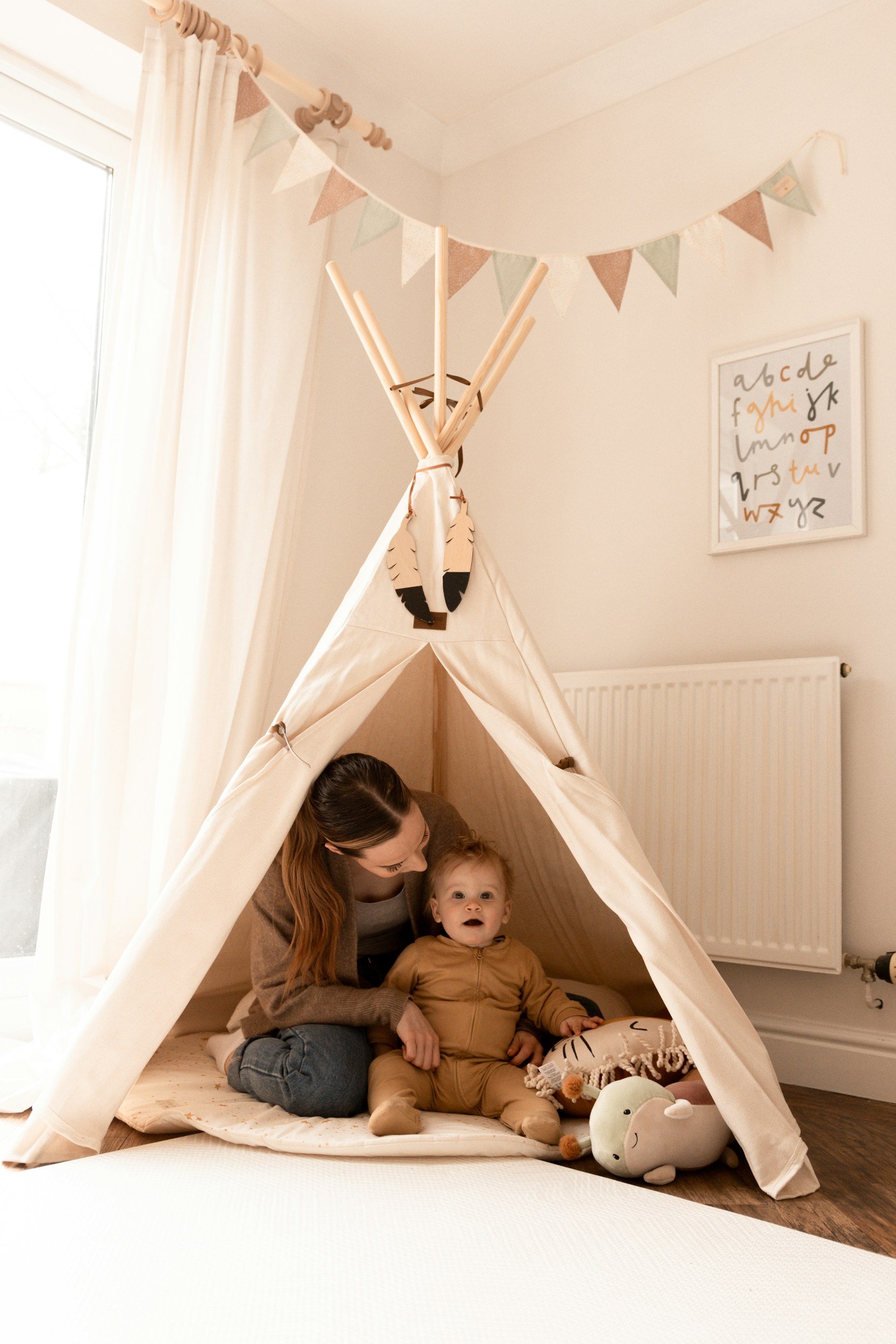 A woman and a young child sitting inside a cream-colored teepee tent in a brightly lit room with white walls, a radiator, and a window with curtains. Decorative plush toys are on the floor.
