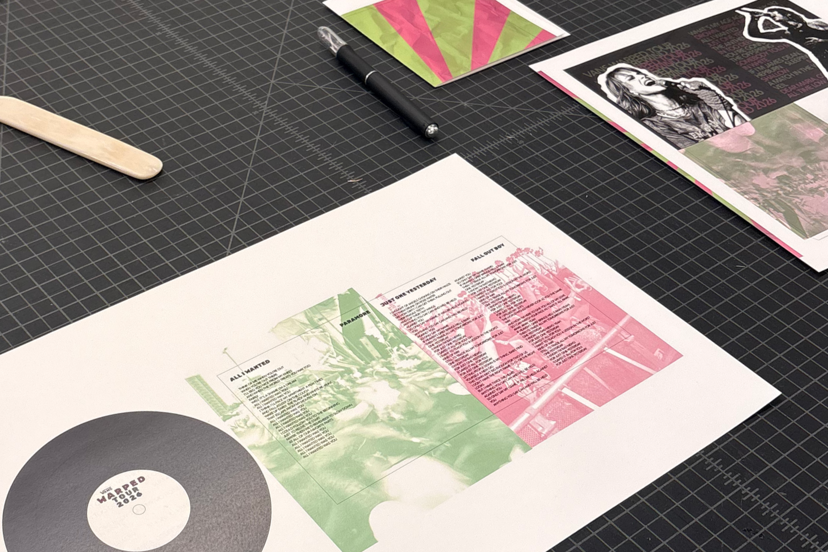A printing workspace with a cutting mat, printed posters, a pen, and a bone folder. The posters feature texts, images, and graphic designs in pink and green.