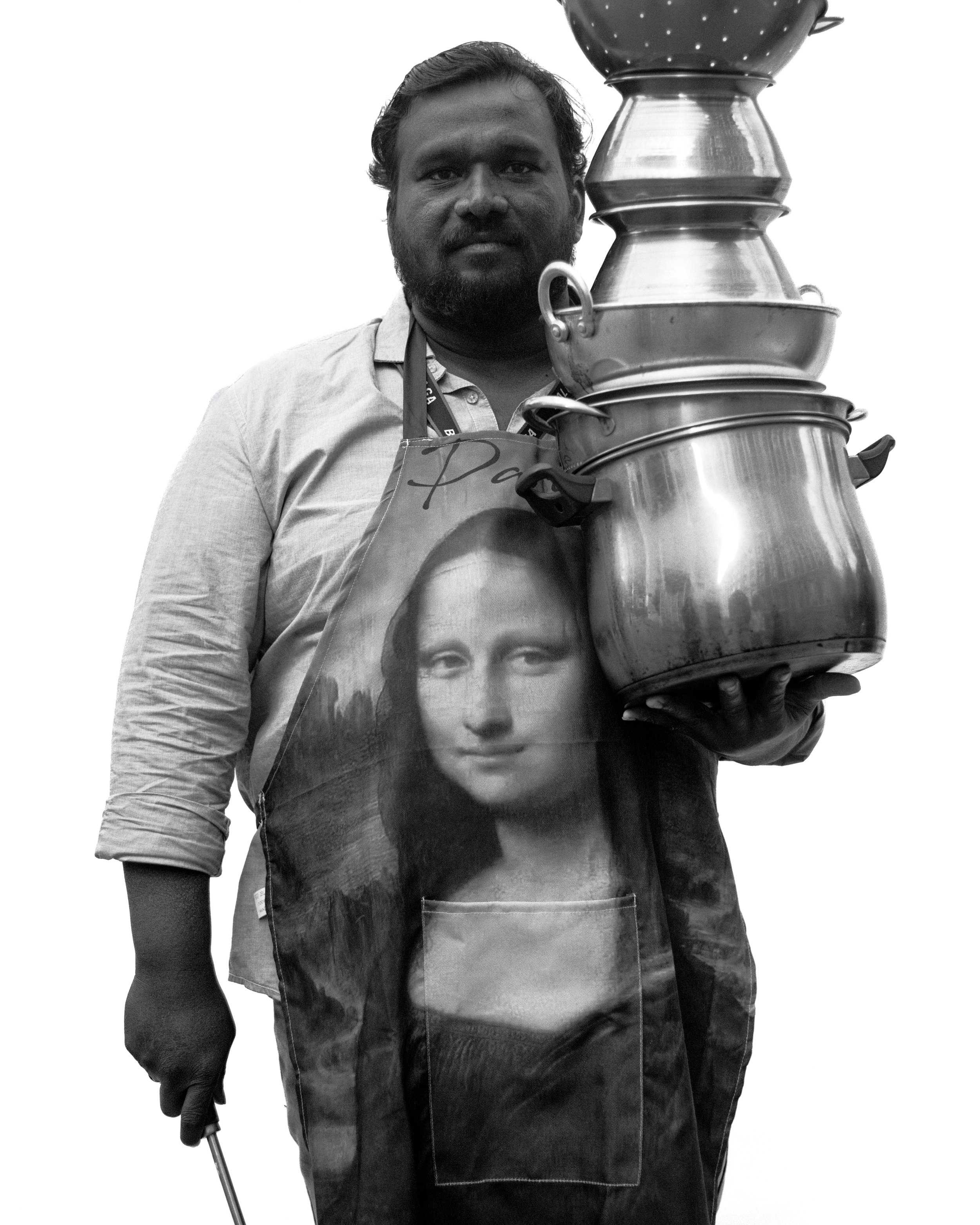 Un homme avec une barbe portant un tablier avec une image de Mona Lisa, tenant une main un objet métallique et une autre une casserole.