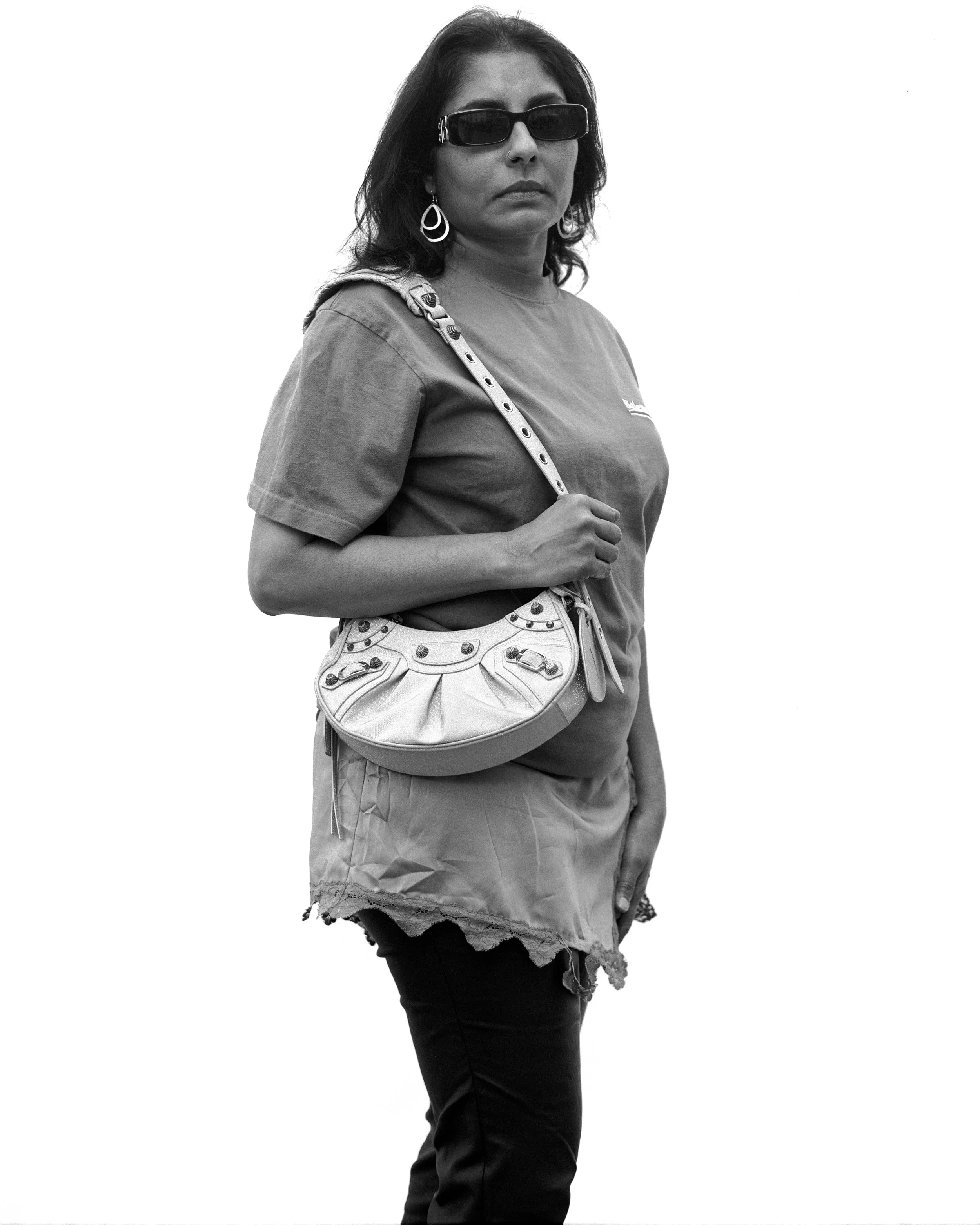 Une femme portant des lunettes de soleil, une chemise décontractée, une jupe déchirée et un sac à bandoulière, posant contre un fond blanc.