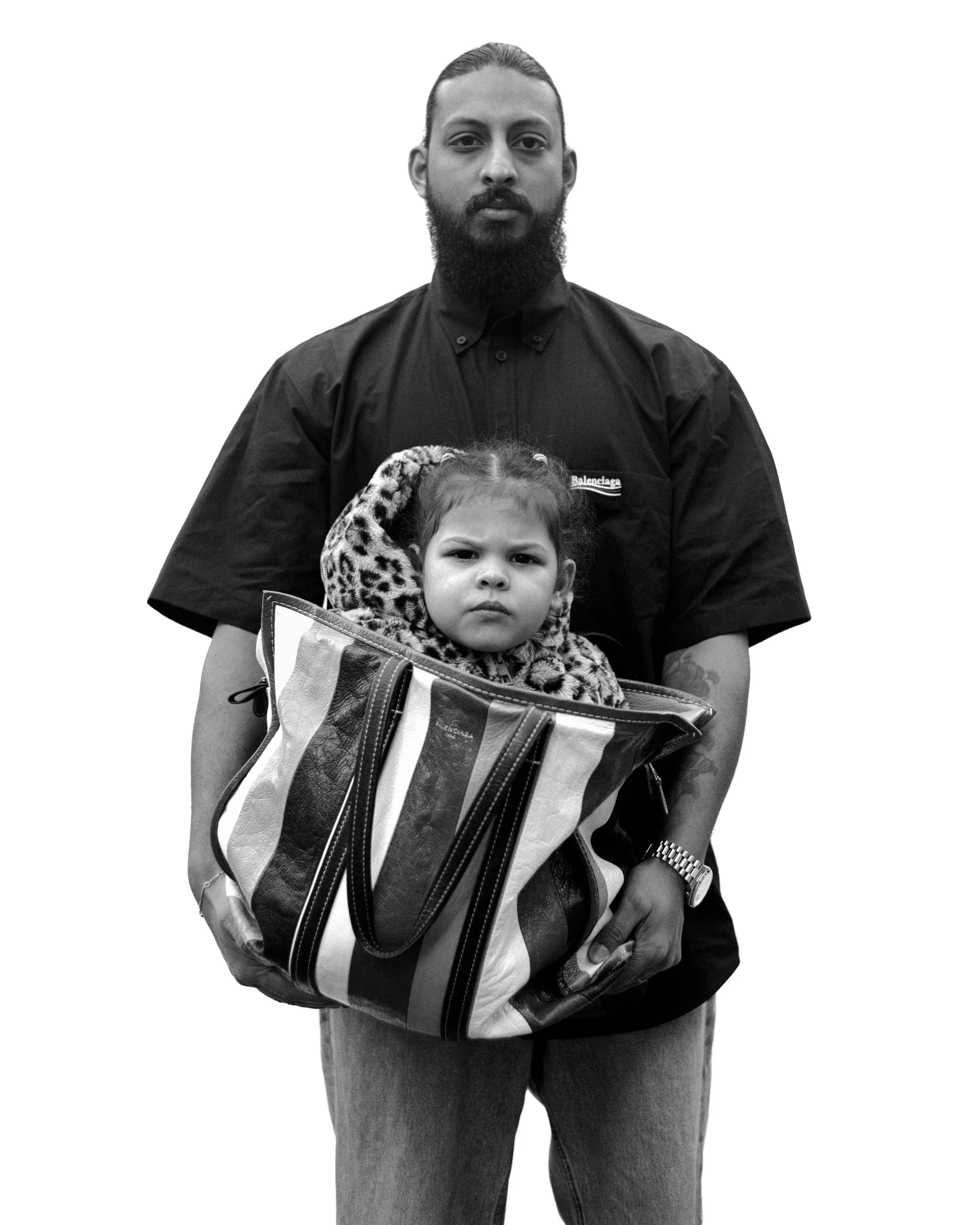 Un homme avec une barbe et une montée de cheveux, portant une chemise noire, tient une jeune fille dans un sac de voyage multicolore. La fille a les cheveux bruns, porte un manteau à motif léopard et regarde sérieusement.