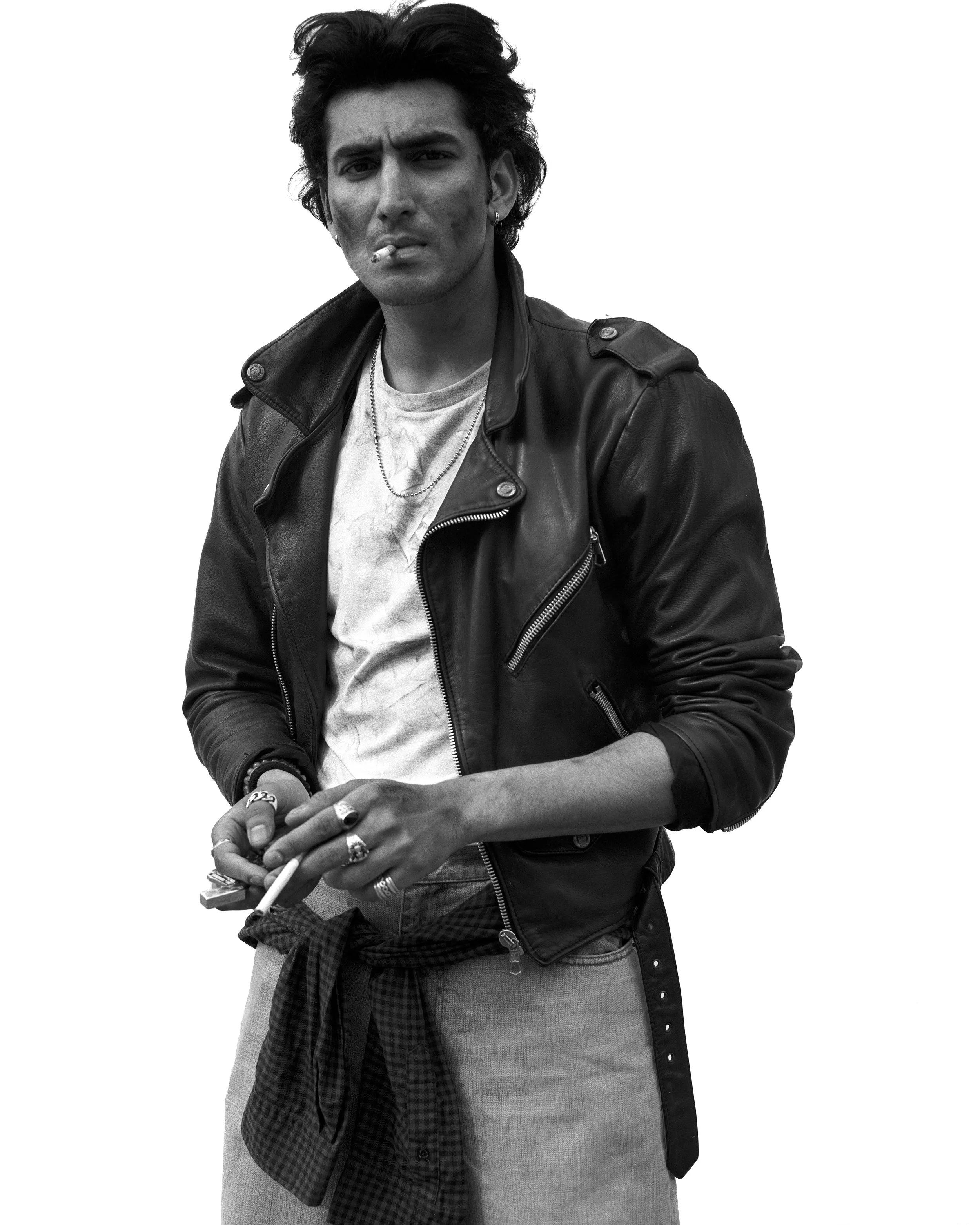 Jeune homme avec des cheveux bouclés, portant une veste en cuir, un t-shirt blanc, plusieurs bagues, un collier, tenant une cigarette, sans yeux ou fond.