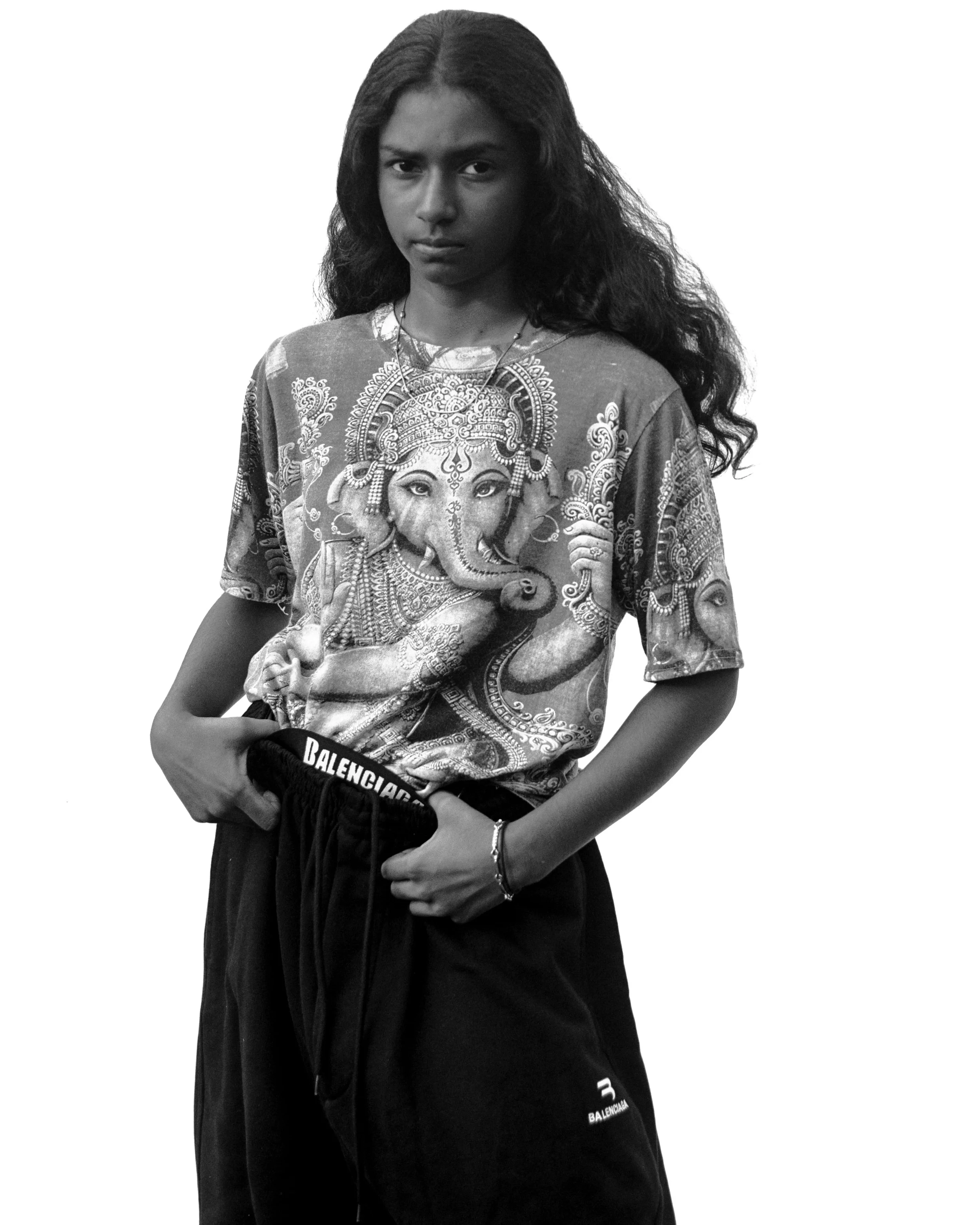 Une jeune femme avec de longs cheveux bouclés, portant un t-shirt avec une image de Ganesha et un pantalon de marque Balenciaga, debout contre un fond blanc.