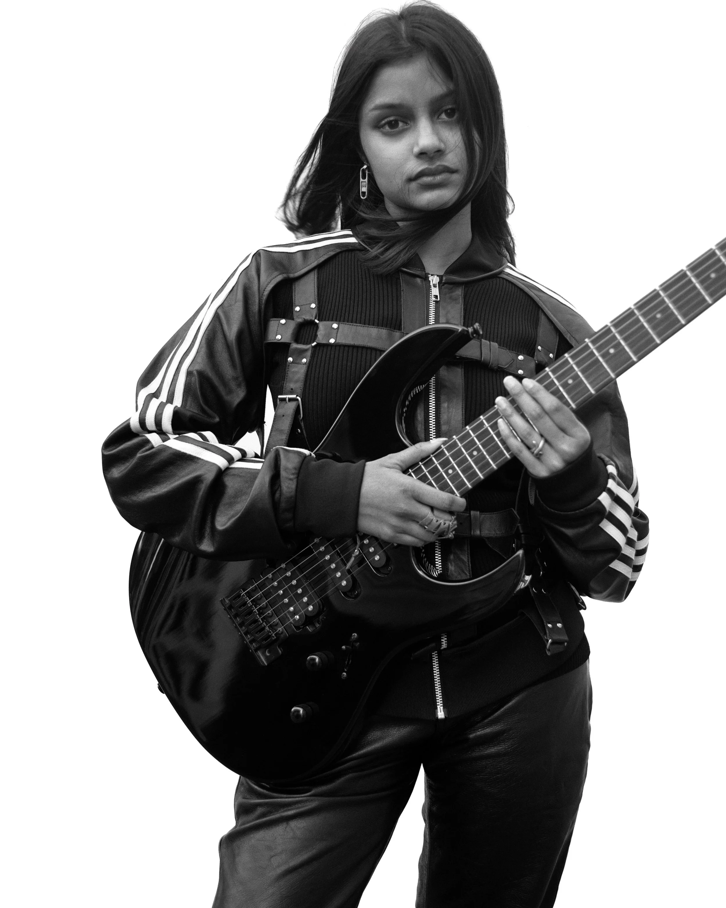 Femme tenant une guitare électrique en noir et blanc, portant une veste en cuir à rayures, avec une expression sérieuse.