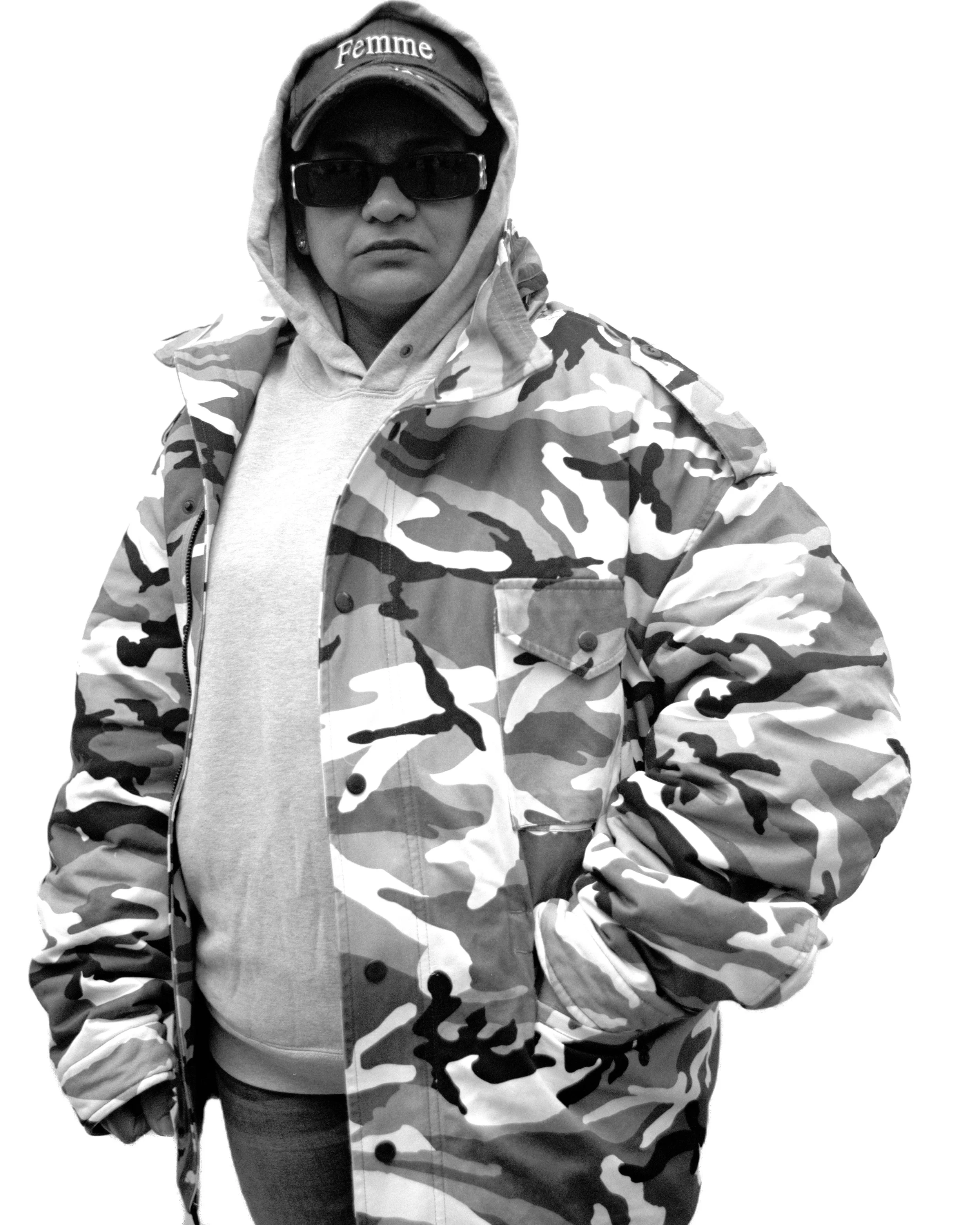 Une personne portant un manteau camouflage, un bonnet avec le mot "Femme", des lunettes de soleil et un sweat à capuche, pose contre un fond blanc.