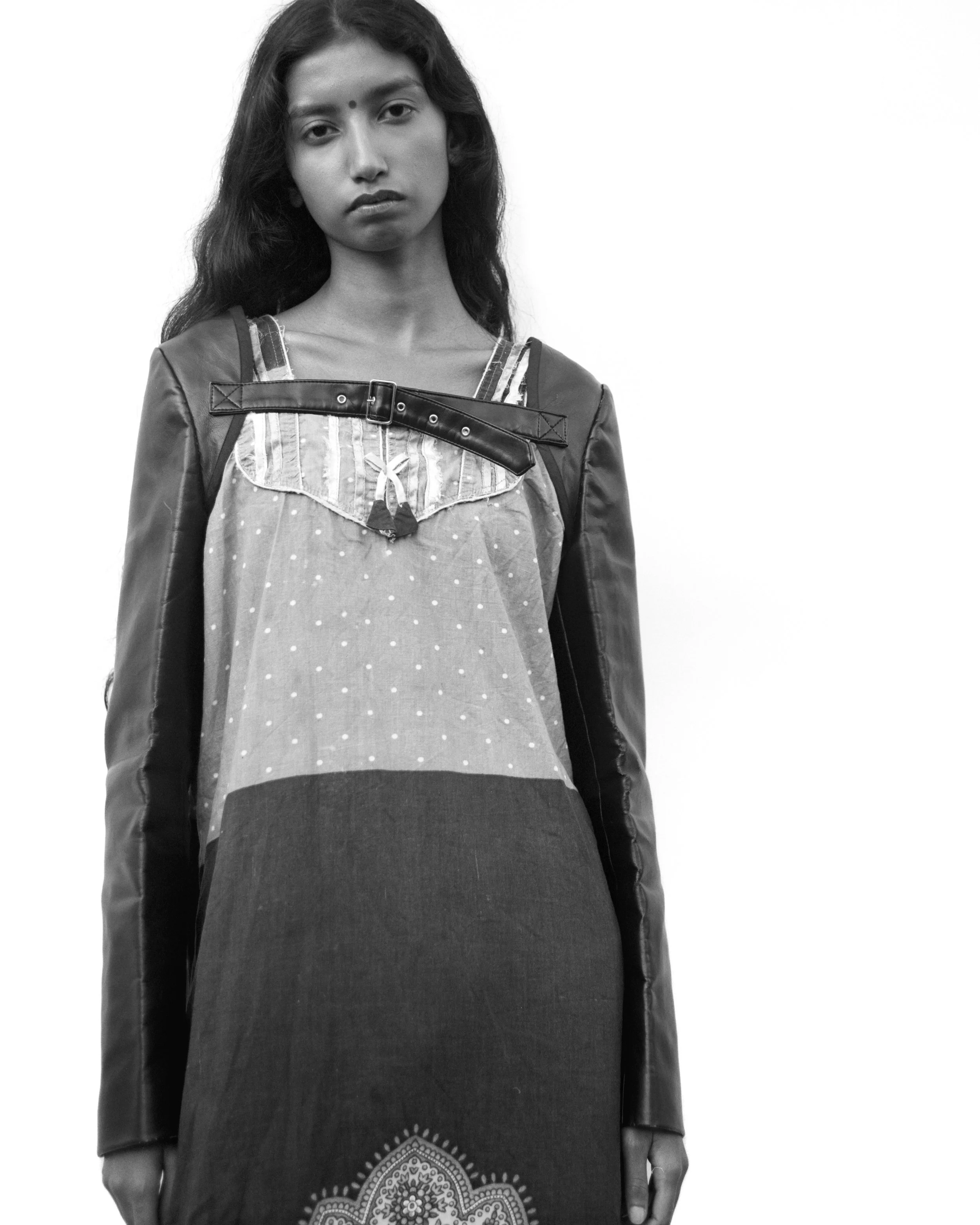 Une jeune femme posant contre un mur blanc, portant une robe à motifs et une veste en cuir.