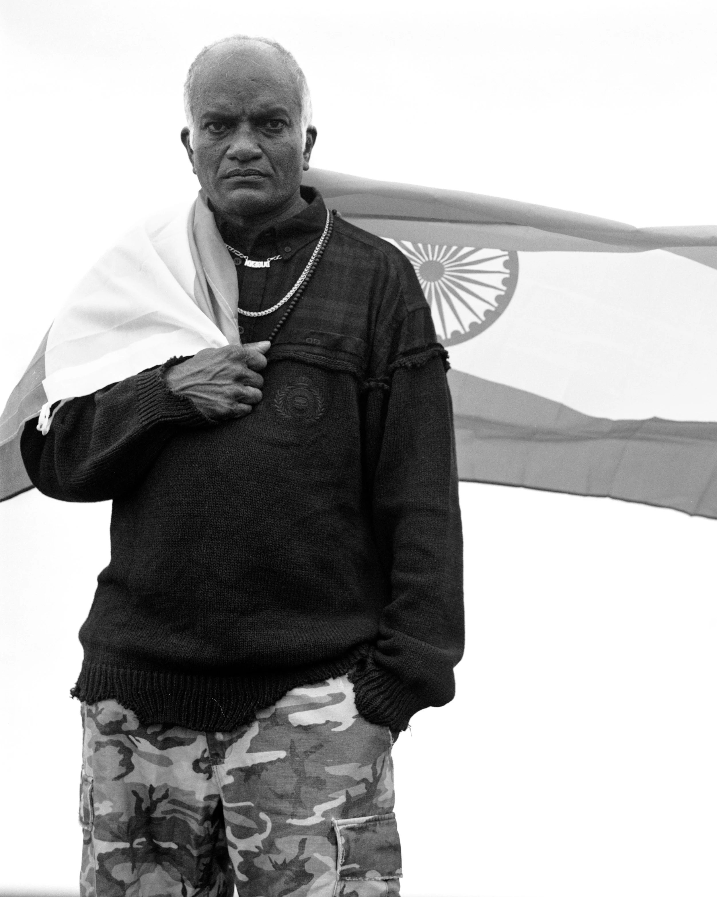 Un homme noir portant un pull noir, des chaînes, et un drapeau indien sur l'épaule, avec un drapeau indien en arrière-plan.
