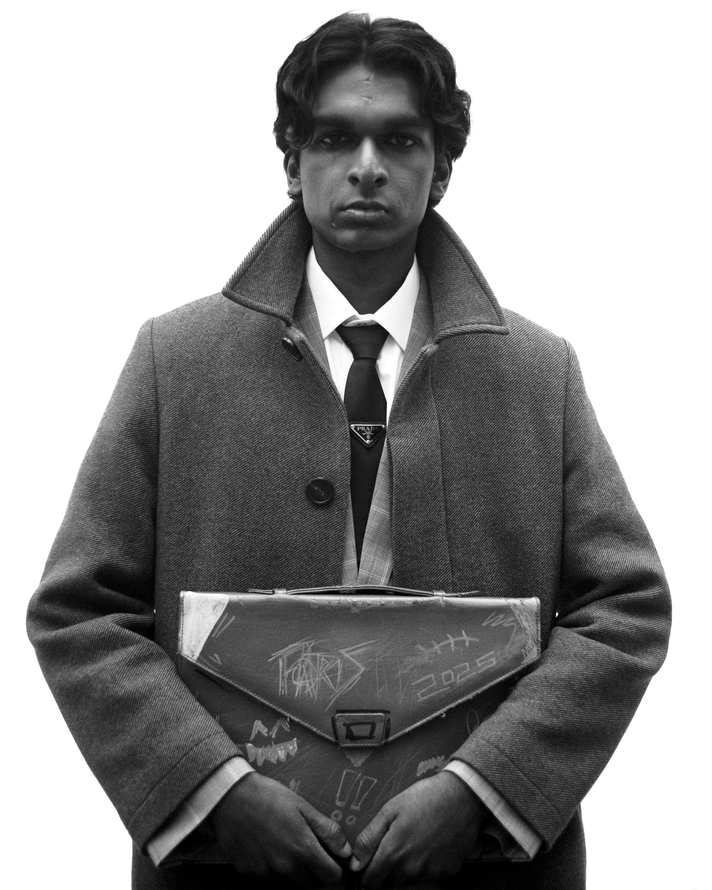 Un homme porte un manteau en laine, une chemise blanche, une cravate avec une broche Prada, et tient un cartable décoré de dessins. Il a des cheveux ondulés et une expression sérieuse sur le visage.