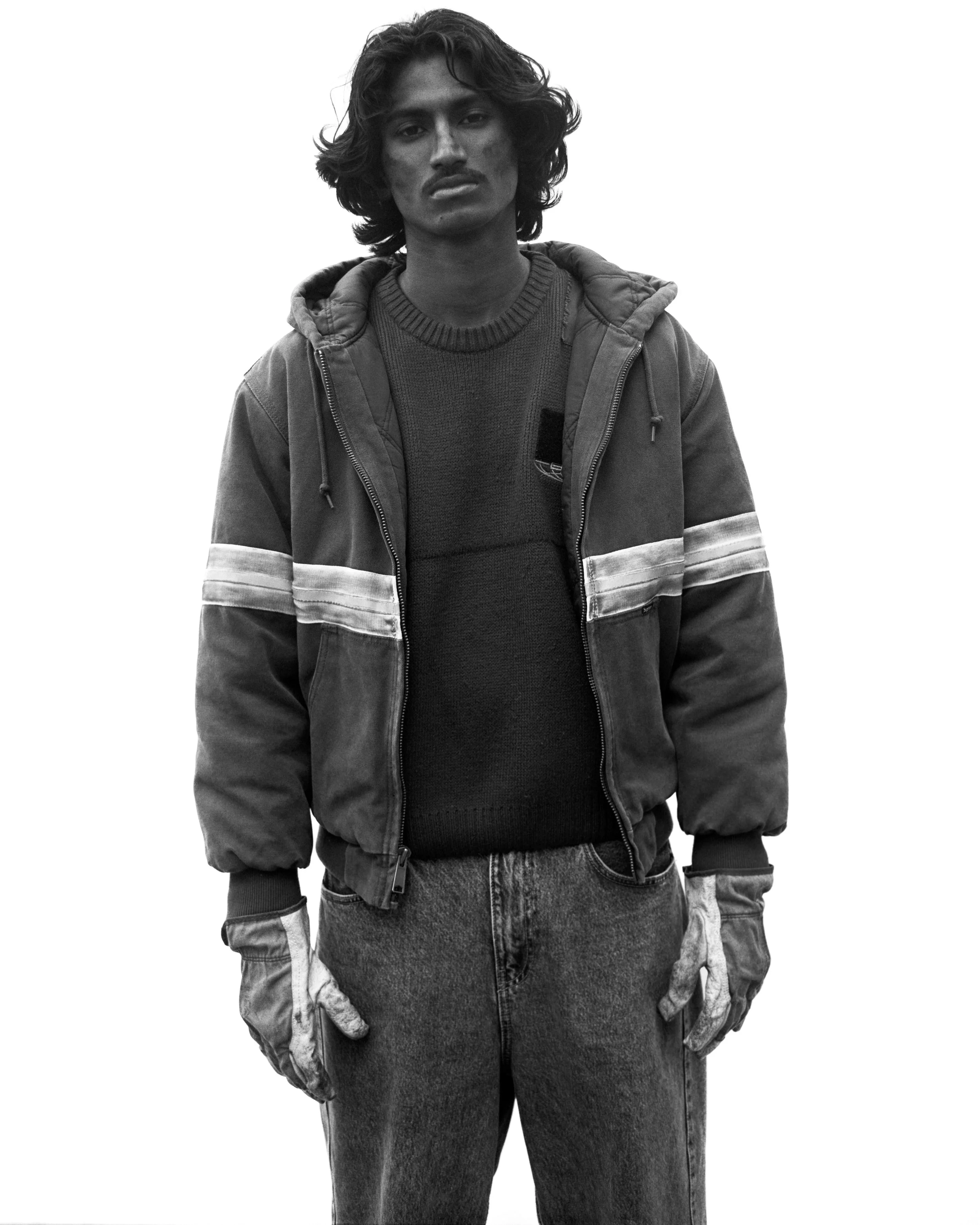 Jeune homme avec cheveux longs, veste d'hiver et gants, debout contre un fond blanc.