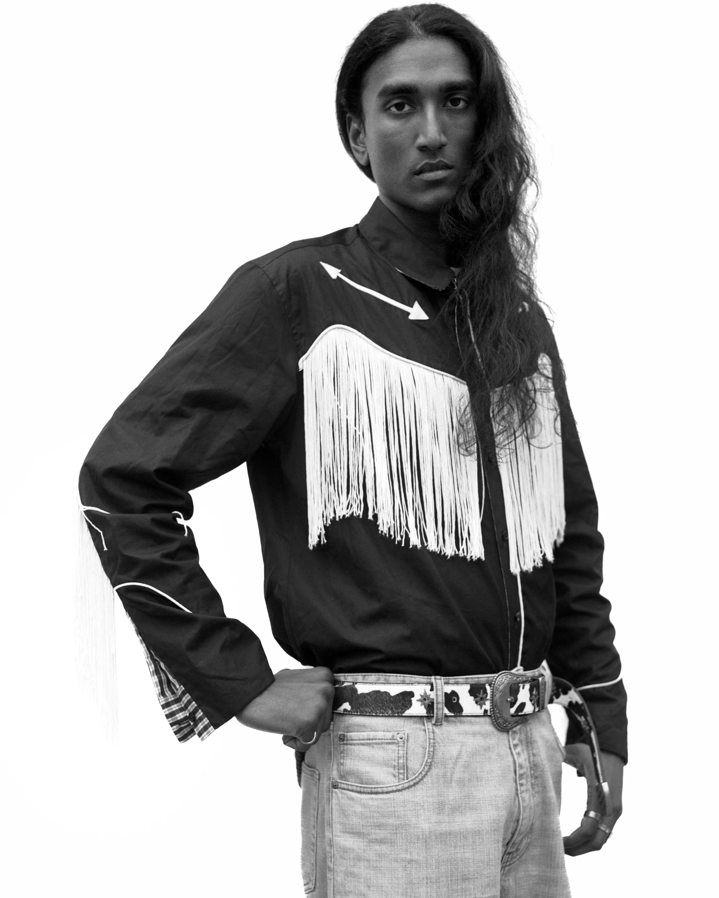 Portrait en noir et blanc d'une femme avec des cheveux longs ondulés portant une chemise à franges et un jean, posant avec la main sur la hanche.