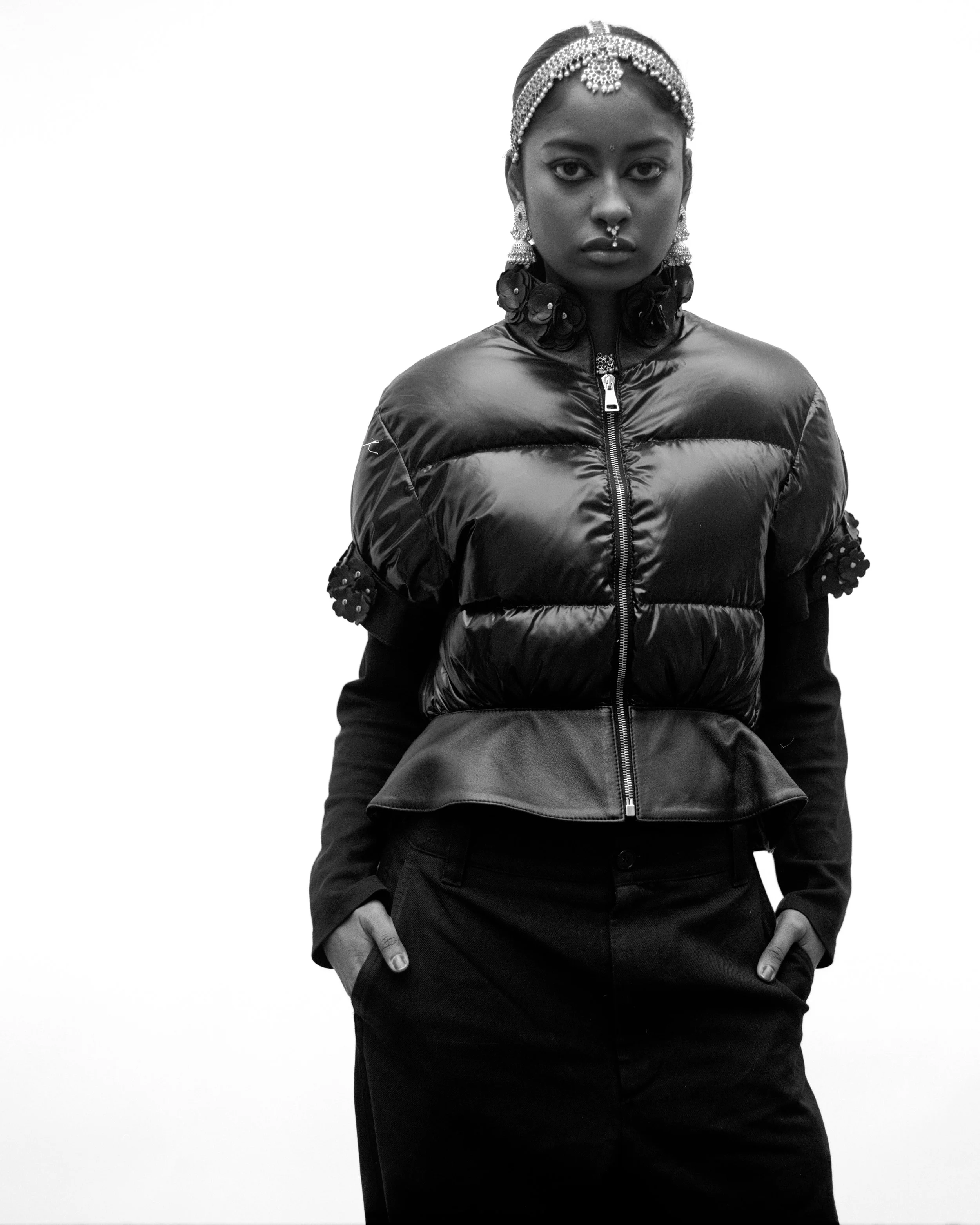 Une femme porte des bijoux ethniques, un tissu en perles sur la tête, une veste épaissie et des vêtements sombres.