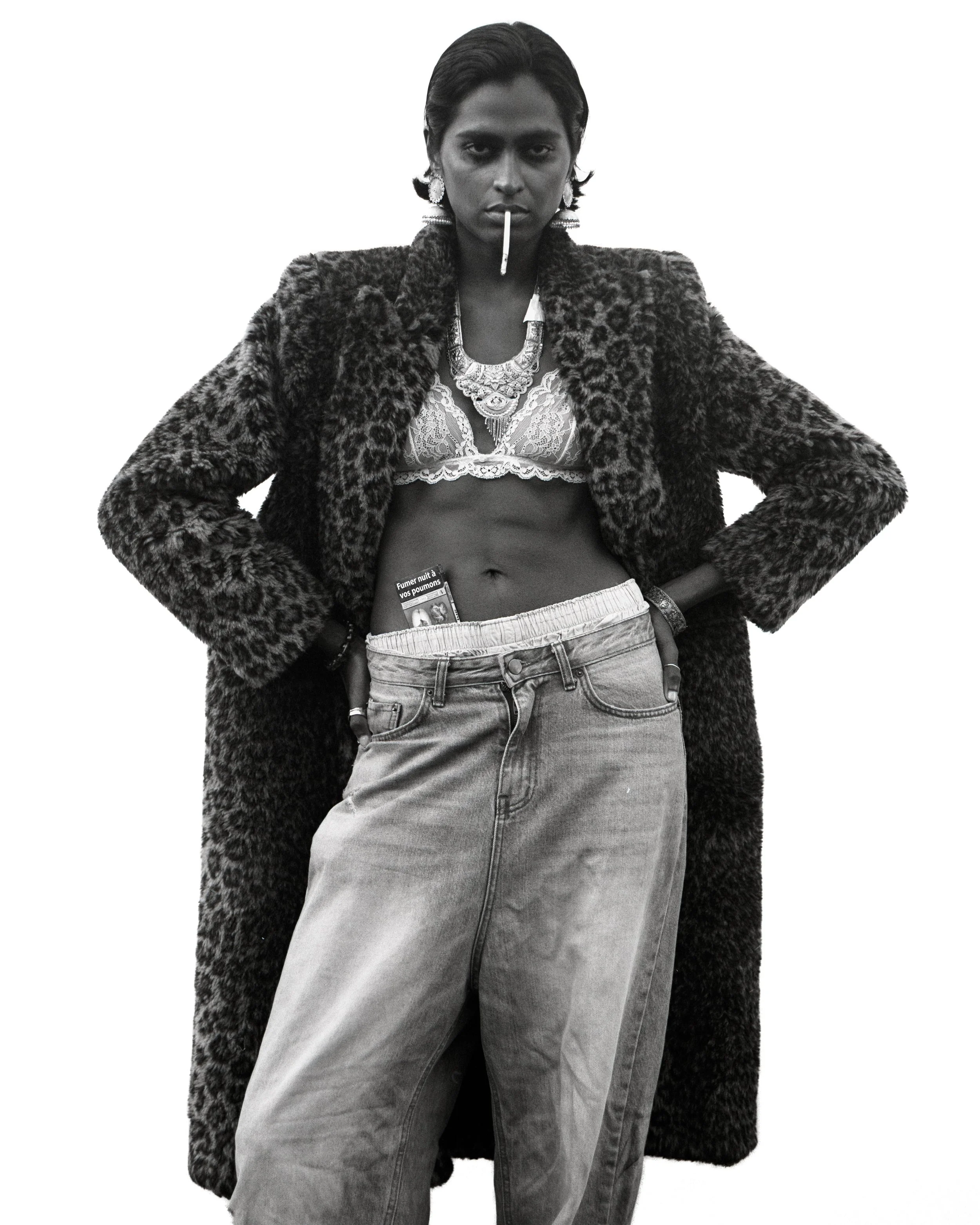 Femme avec des vêtements décontractés, portant un manteau en fausse fourrure, un top en dentelle, un jean, et un cigare dans la bouche, pose de manière confiante.