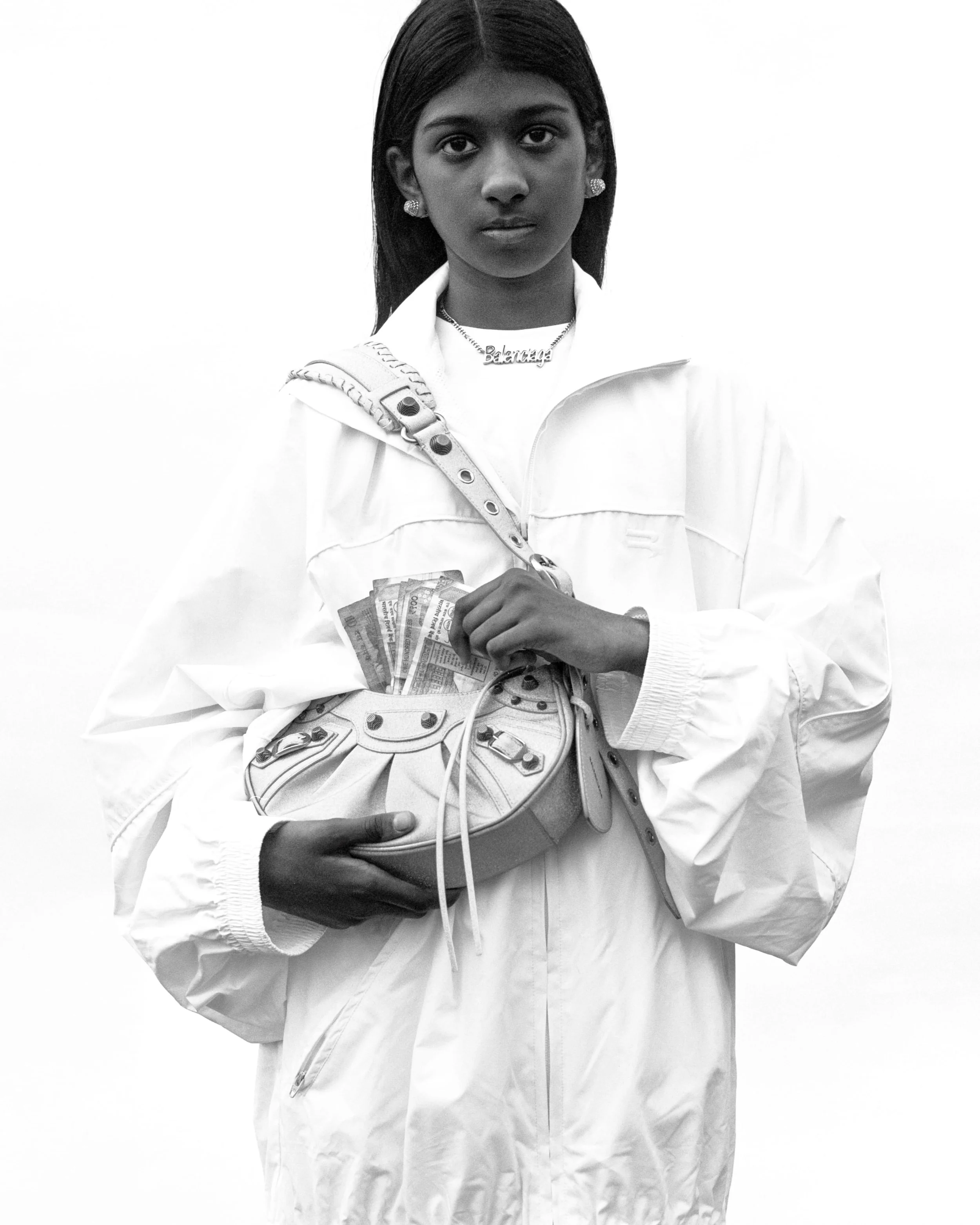 Une femme noire portant une veste blanche, des boucles d'oreilles, un collier avec le mot 'Balance' et tenant un sac à main contenant de l'argent et des billets.