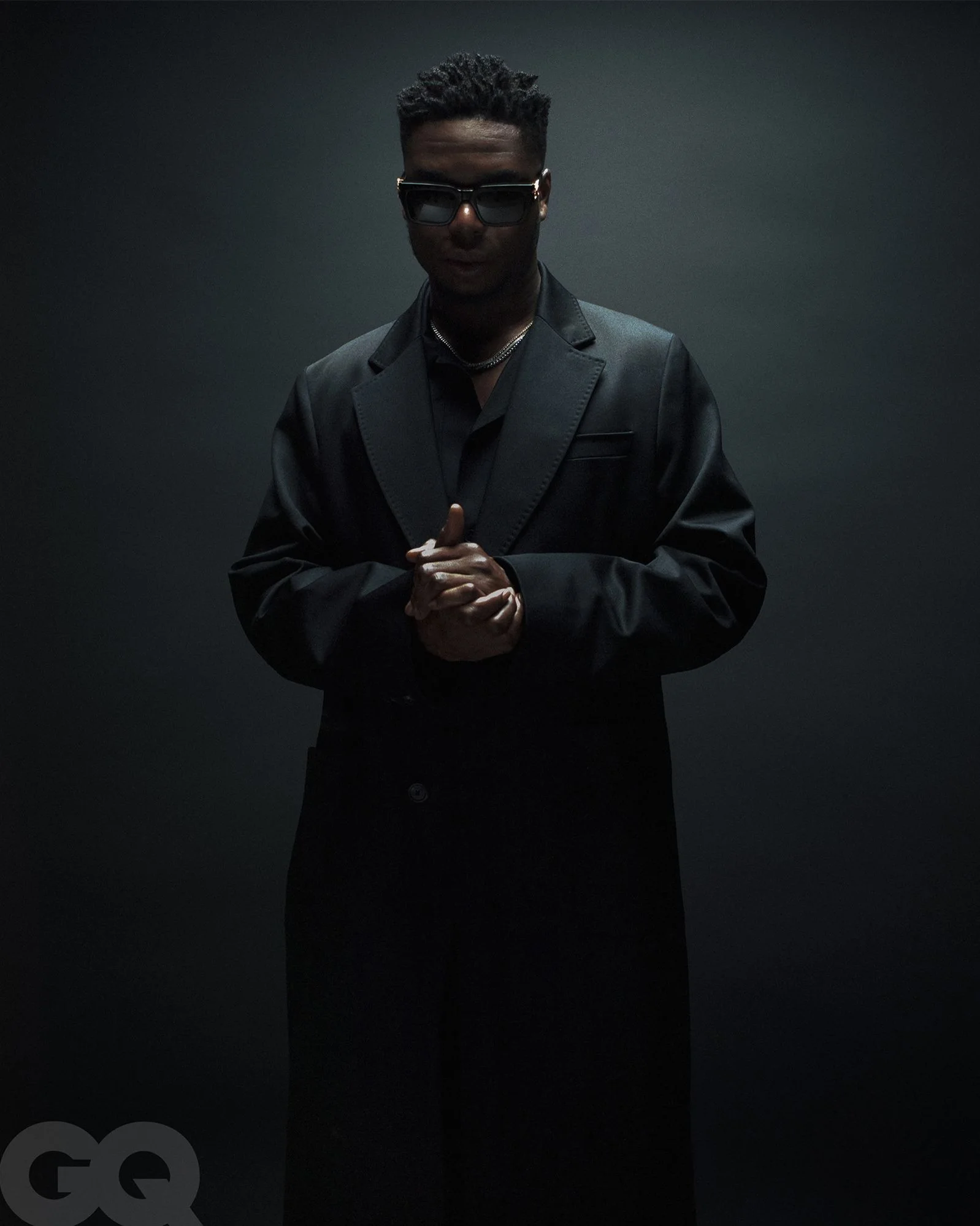 Un homme habillé en noir, portant des lunettes de soleil et une chaîne, se tient contre un fond sombre.