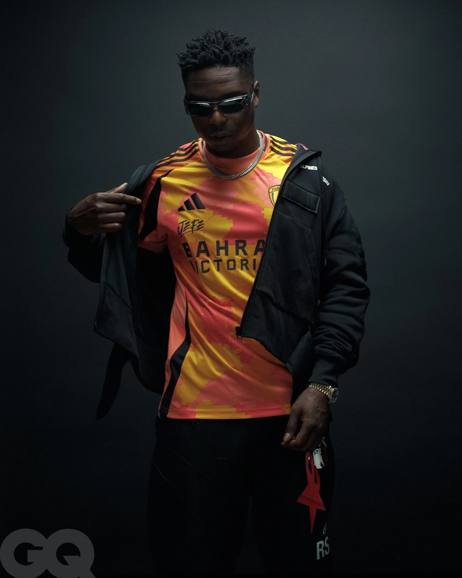 Un homme noir portant des lunettes de soleil, un maillot de football aux couleurs orange et jaune avec le logo d'une équipe, une veste noire ouverte, une montre en or, et un sac à dos noir, posé contre un fond sombre.