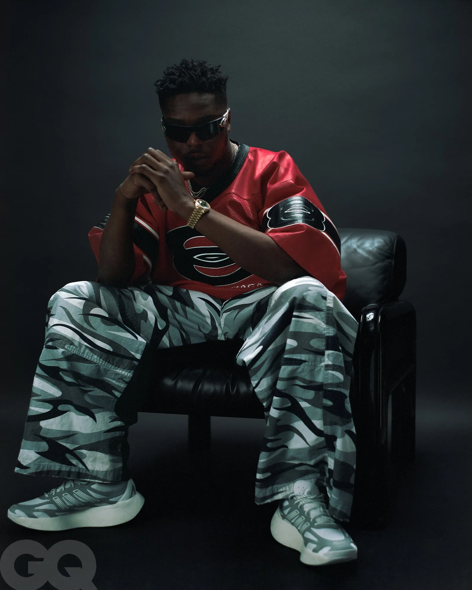 Un homme assis dans un fauteuil noir, portant des vêtements de style urbain avec une casquette et des lunettes de soleil, pose de manière confiante.