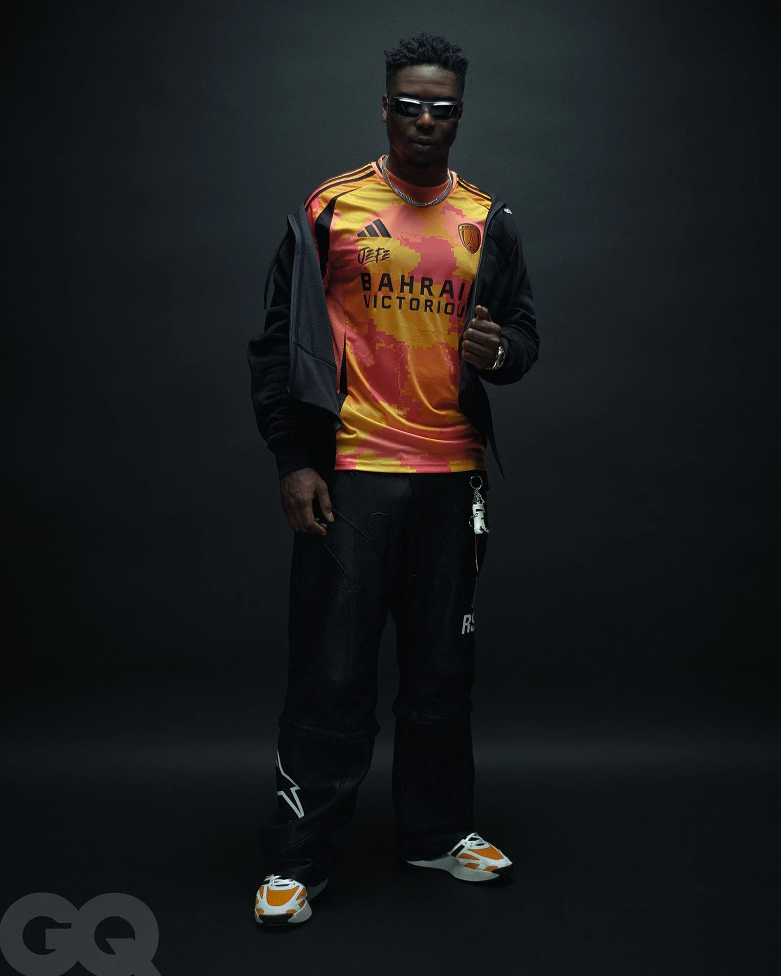 Un homme portant un maillot de football de l'équipe Bahrain Victory, un sweat noir, des pantalons noirs et des chaussures de sport blanches et orange, pose devant un fond noir.