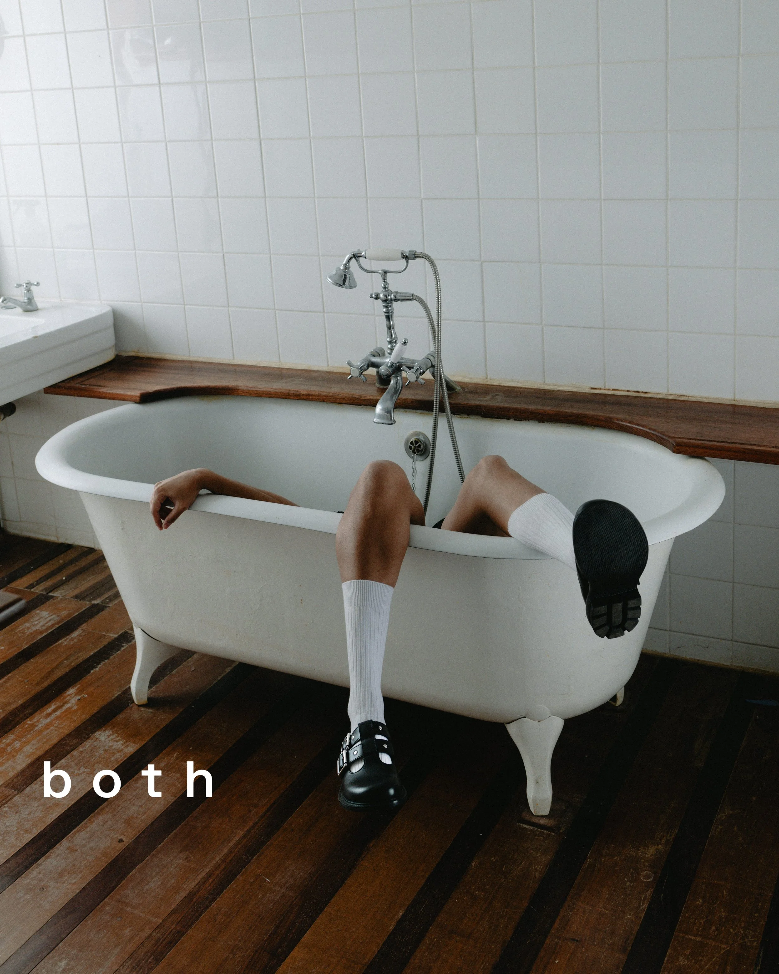 Une personne avec des jambes dénudées dans une baignoire ancienne en salle de bain, portant des chaussettes longues et des chaussures noires, positionnée de façon étrange, avec le mot 'both' écrit en bas à gauche.