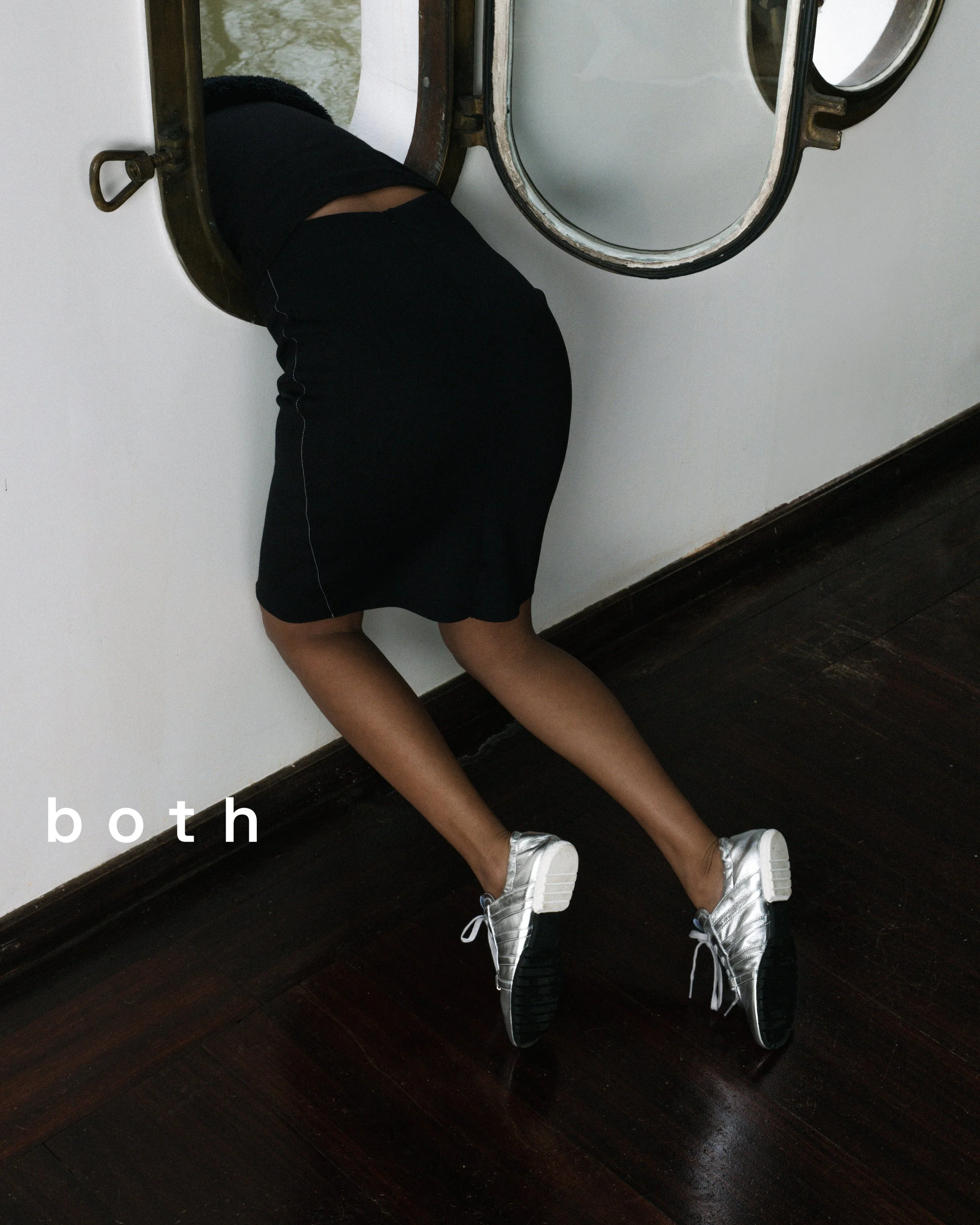 Une personne au corps féminin portant une jupe noire et des chaussures argentées, est allongée dans une machine à laver. La personne a une peau foncée et la scène est vue de côté avec un plan rapproché.