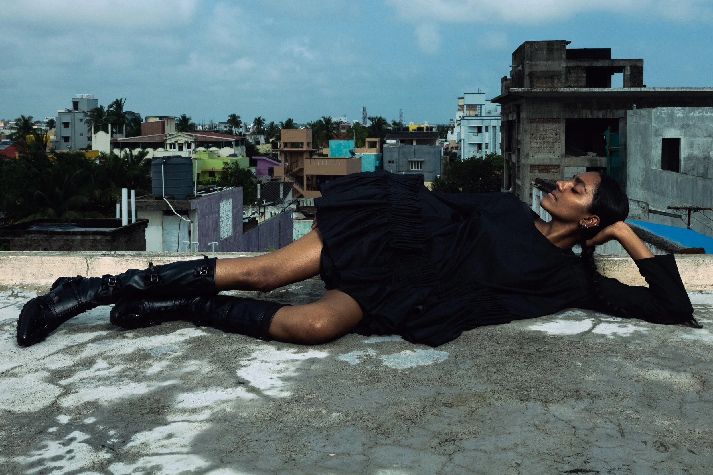 Femme allongée sur un toit en ville, portant une robe noire et des bottes noirs, regardant vers le ciel avec un fond de bâtiments résidentiels et de nuages.