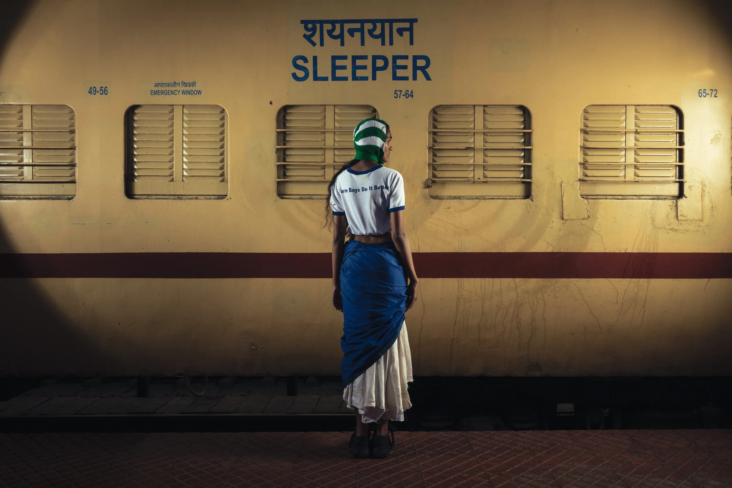 Une femme vêtue d'une chemise blanche avec un texte en anglais et d'un sari bleu, portant un casque vert et blanc, se tient devant un train jaune à la station de train. Le train a des numéros de compartiment, des fenêtres avec des grilles et un panne