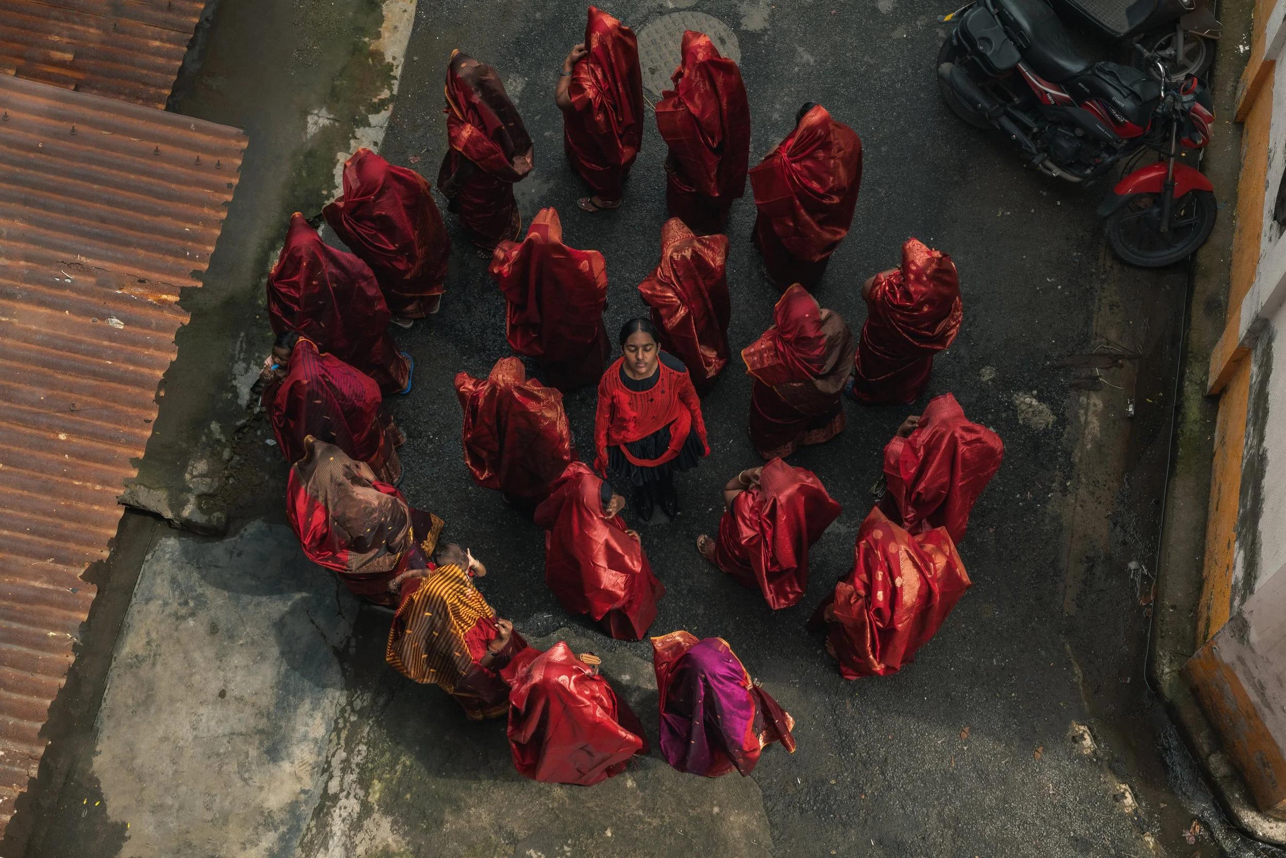 Groupe de femmes portant des vêtements traditionnels rouges, vue du dessus, dans une cour extérieure avec motos et murs.