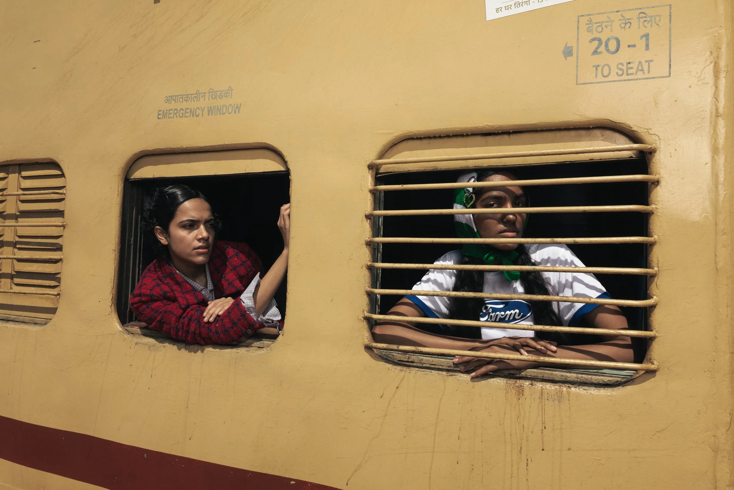Deux jeunes filles regardent par des fenêtres d'un train jaune avec des barres de sécurité. La fille à gauche porte un vêtement rouge et noir, celle à droite porte un t-shirt blanc avec des détails bleus et un foulard vert autour du cou.