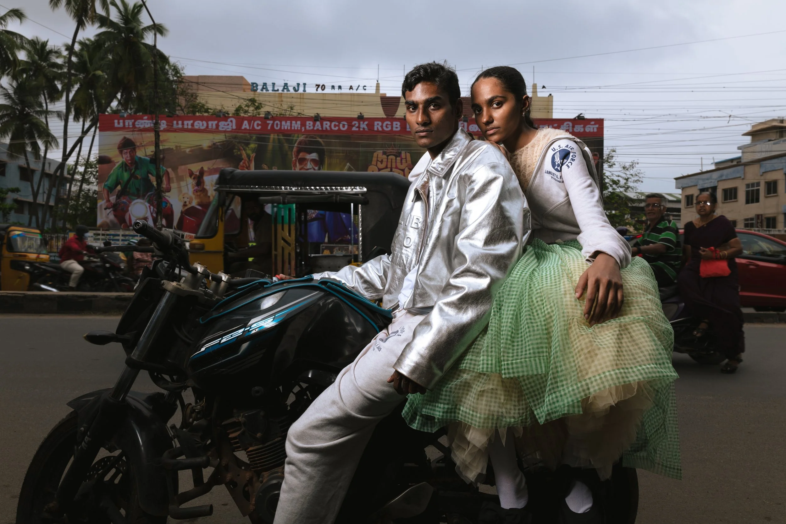 Deux jeunes adultes, un homme et une femme, assis sur une moto dans une rue animée en Inde, avec des bâtiments, des arbres et des passants en arrière-plan.