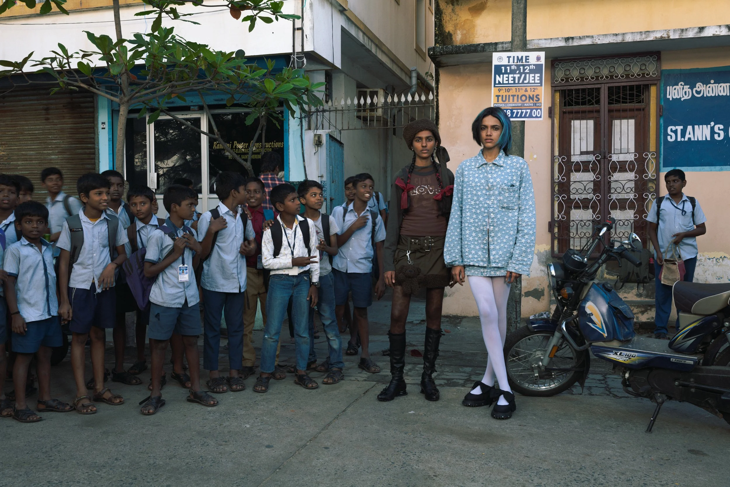 Un groupe d'enfants en uniforme scolaire attend dehors, avec deux jeunes femmes au centre, l'une portant une robe noire et l'autre un vêtement à rayures, devant un bâtiment avec une moto à droite, à l'heure du matin.
