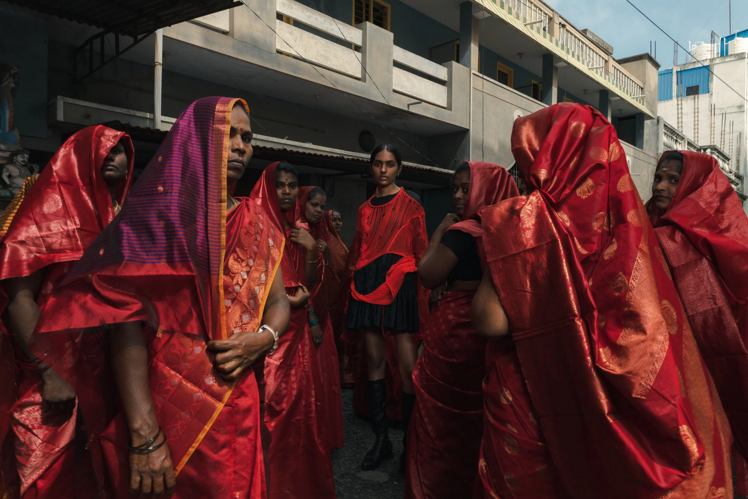 Un groupe de femmes vêtues de saris rouges, certaines avec la tête couverte, se tiennent sur une rue urbaine. Au centre, une jeune femme en robe noire et rouge, portant des bottes, semble être la focalisation de la scène.