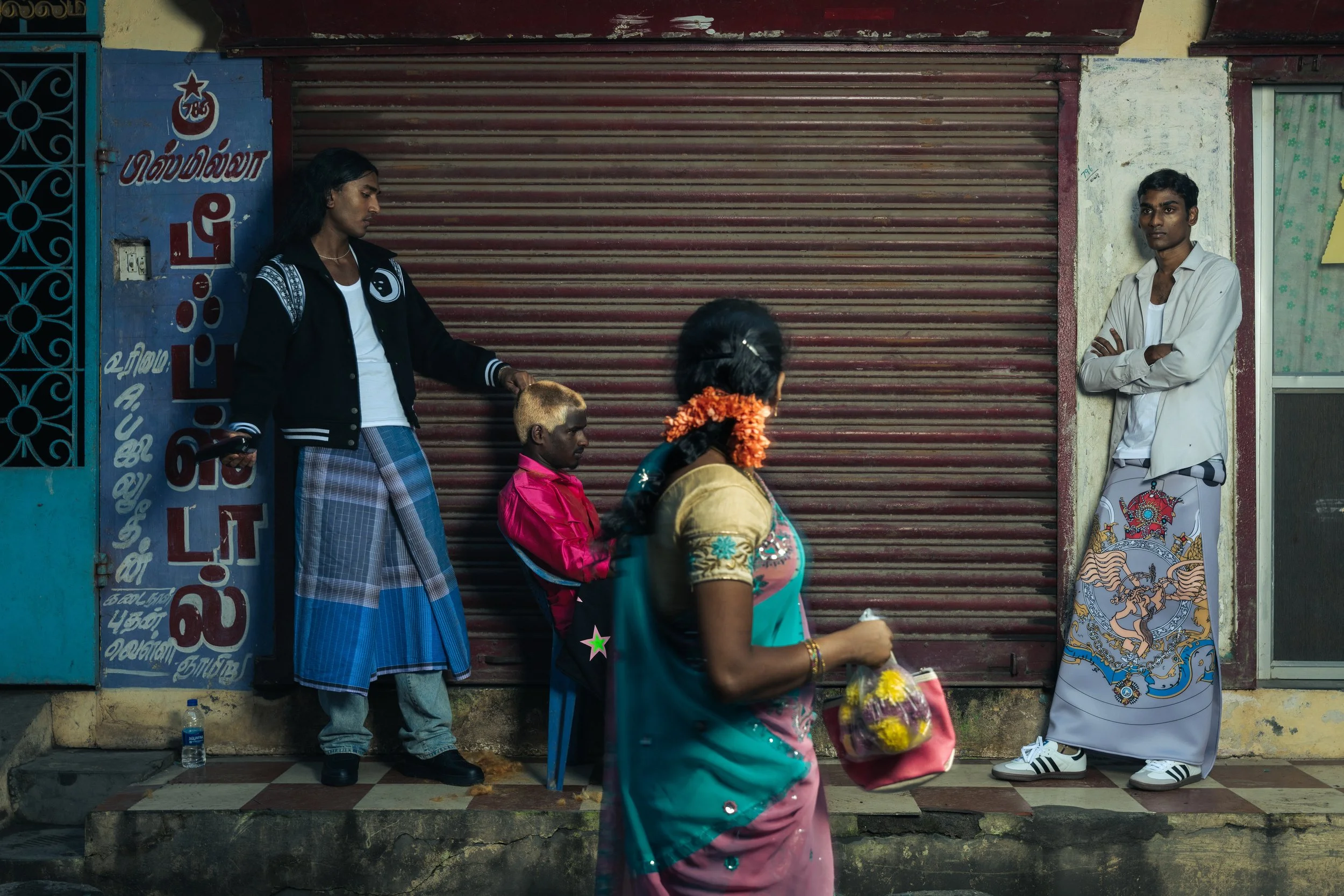 Trois personnes et un enfant se tiennent devant une boutique aux volets métalliques dans une rue. La personne à gauche porte une veste noire, un sari bleu, et regarde un enfant en uniforme rose. La personne à droite porte une chemise blanche et une r