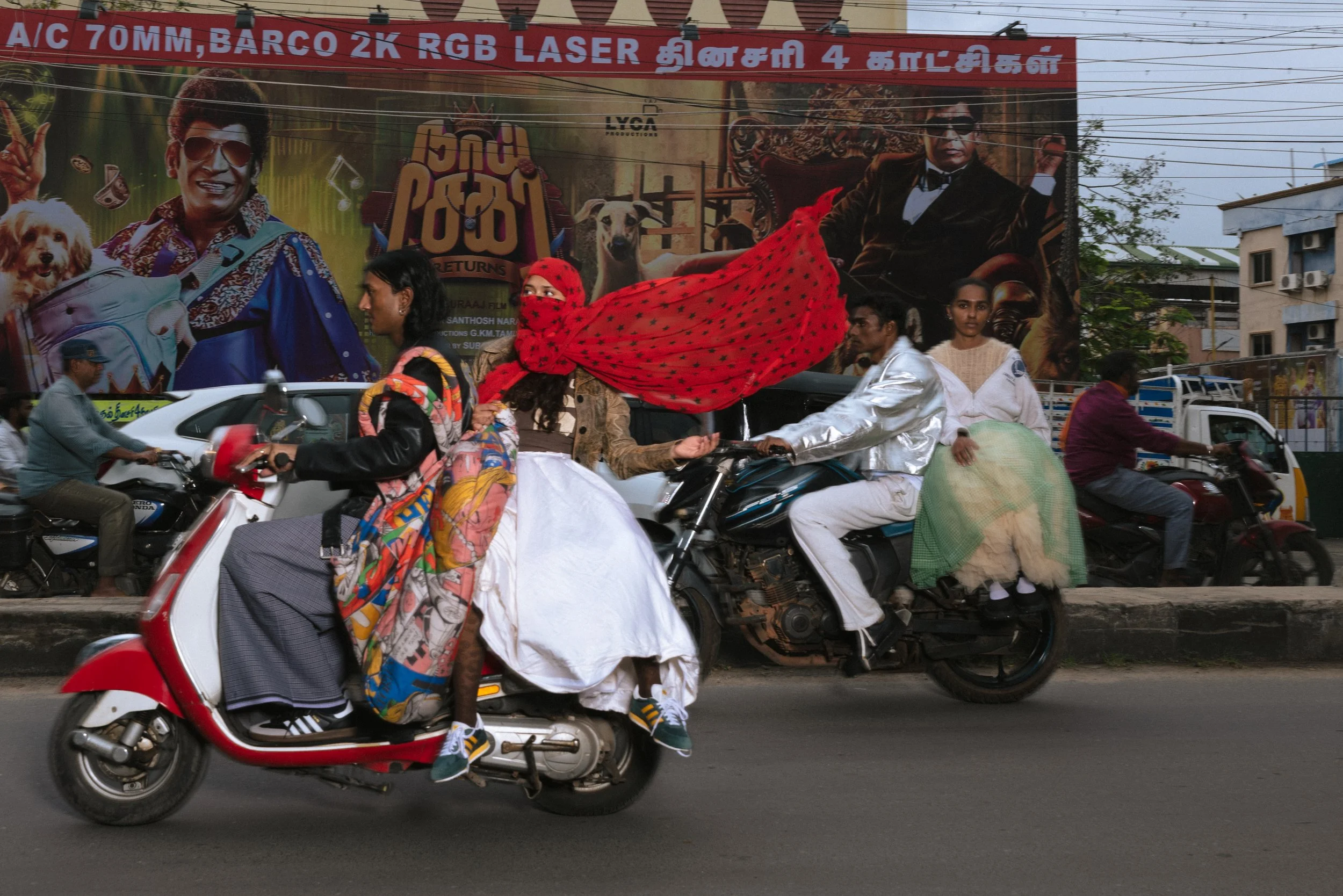 Des personnes en costumes traditionnels indiens conduisent des motos sur la route, avec un grand panneau publicitaire en arrière-plan.