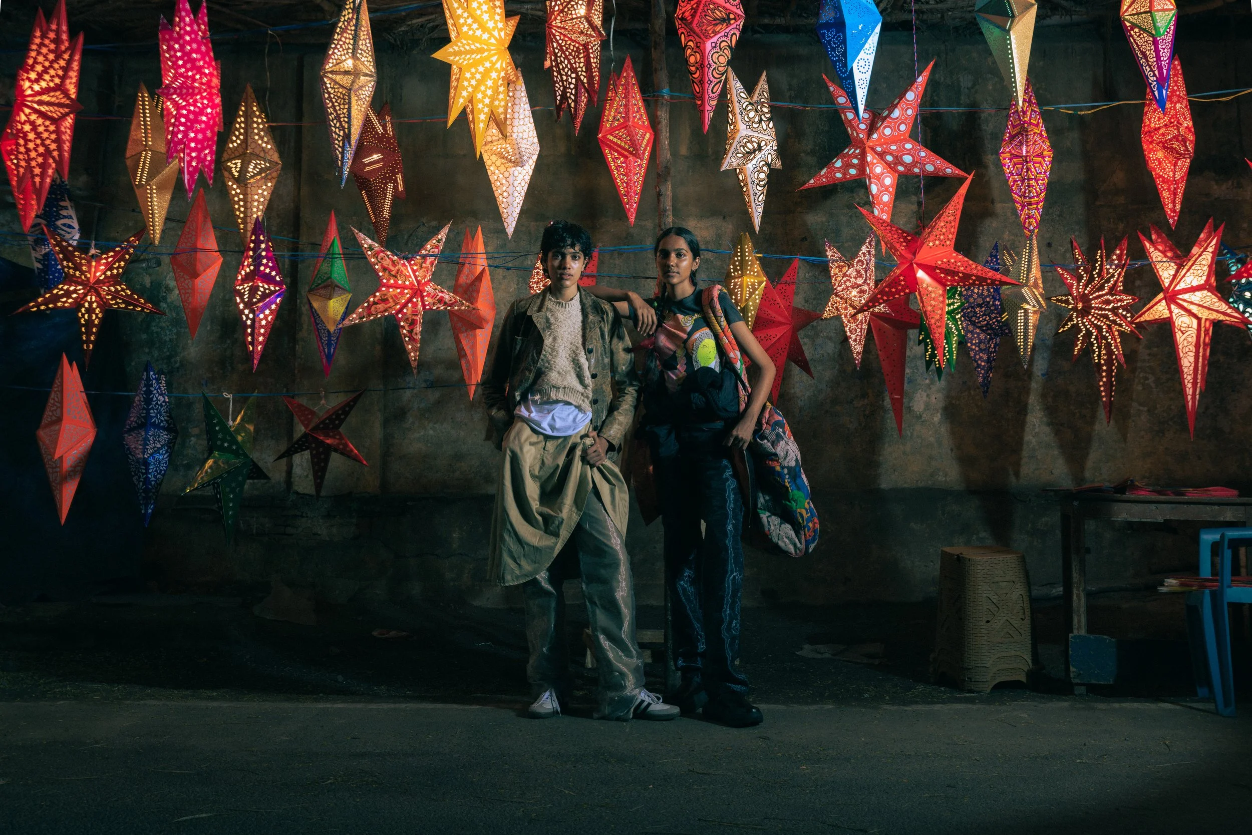 Deux jeunes filles posant devant un mur décoré de lanternes étoilées colorées suspendues. L'une porte un manteau kaki et une jupe beige, l'autre porte un t-shirt imprimé et un sac à dos avec un manteau porté en bandoulière.