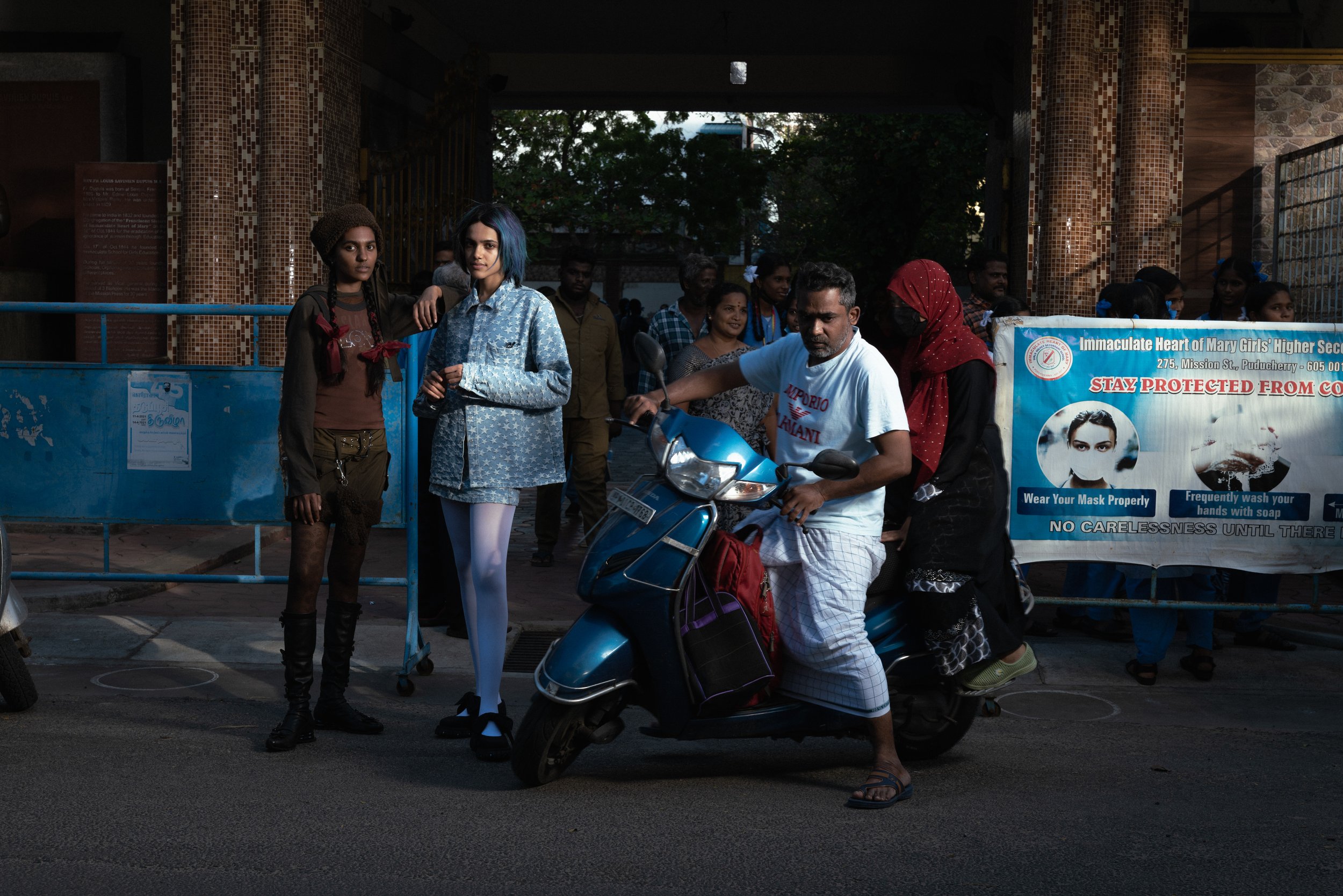 Groupe de personnes devant un bâtiment avec un panneau de sensibilisation à la sécurité sanitaire. Un homme en scooter, une femme avec un masque noir et un sac, et deux jeunes femmes posant pour la photo.