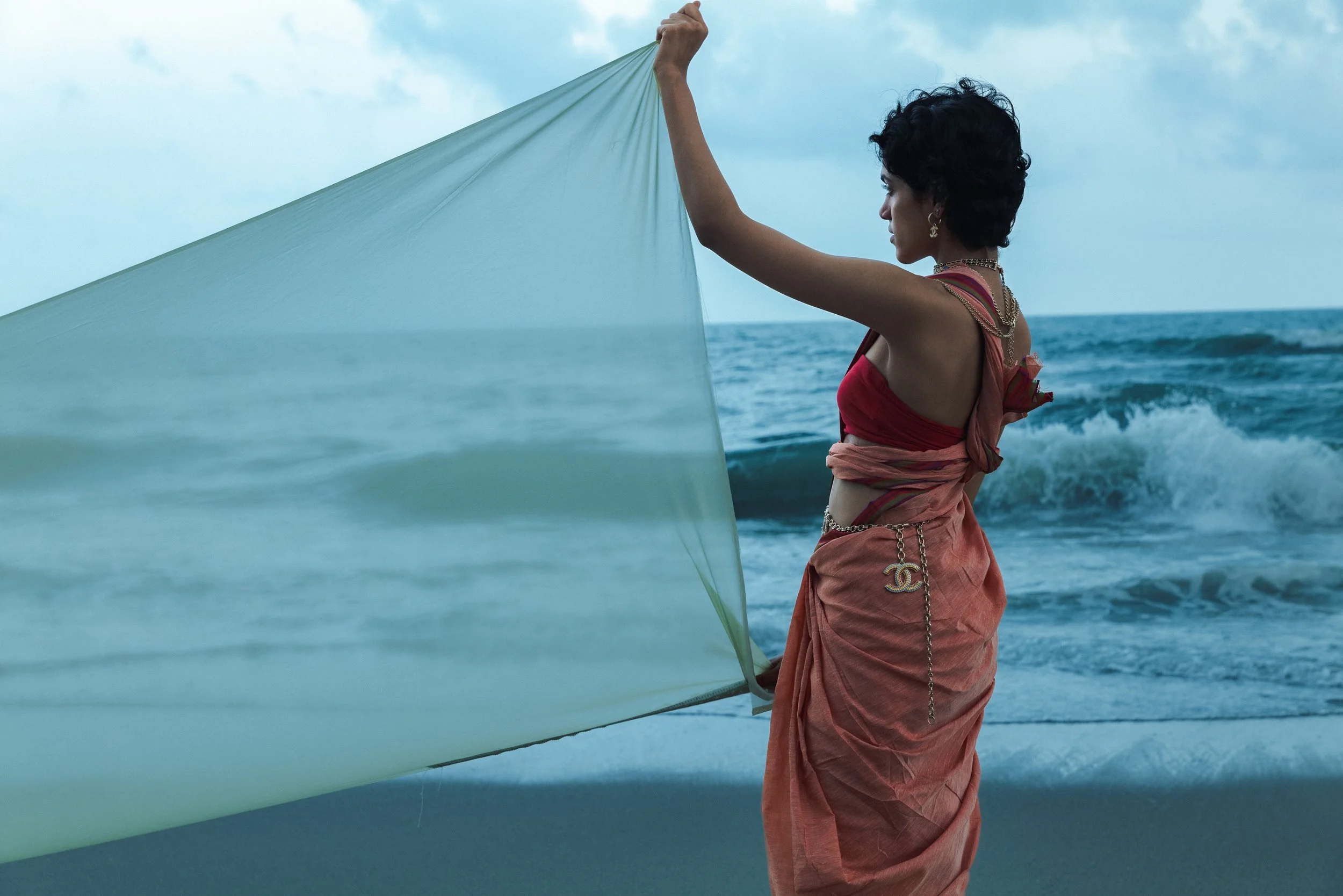 Une femme habillée en tenue de style sarouel, tenant un drapeau blanc au bord de la mer, avec des vagues en arrière-plan, sous un ciel nuageux.