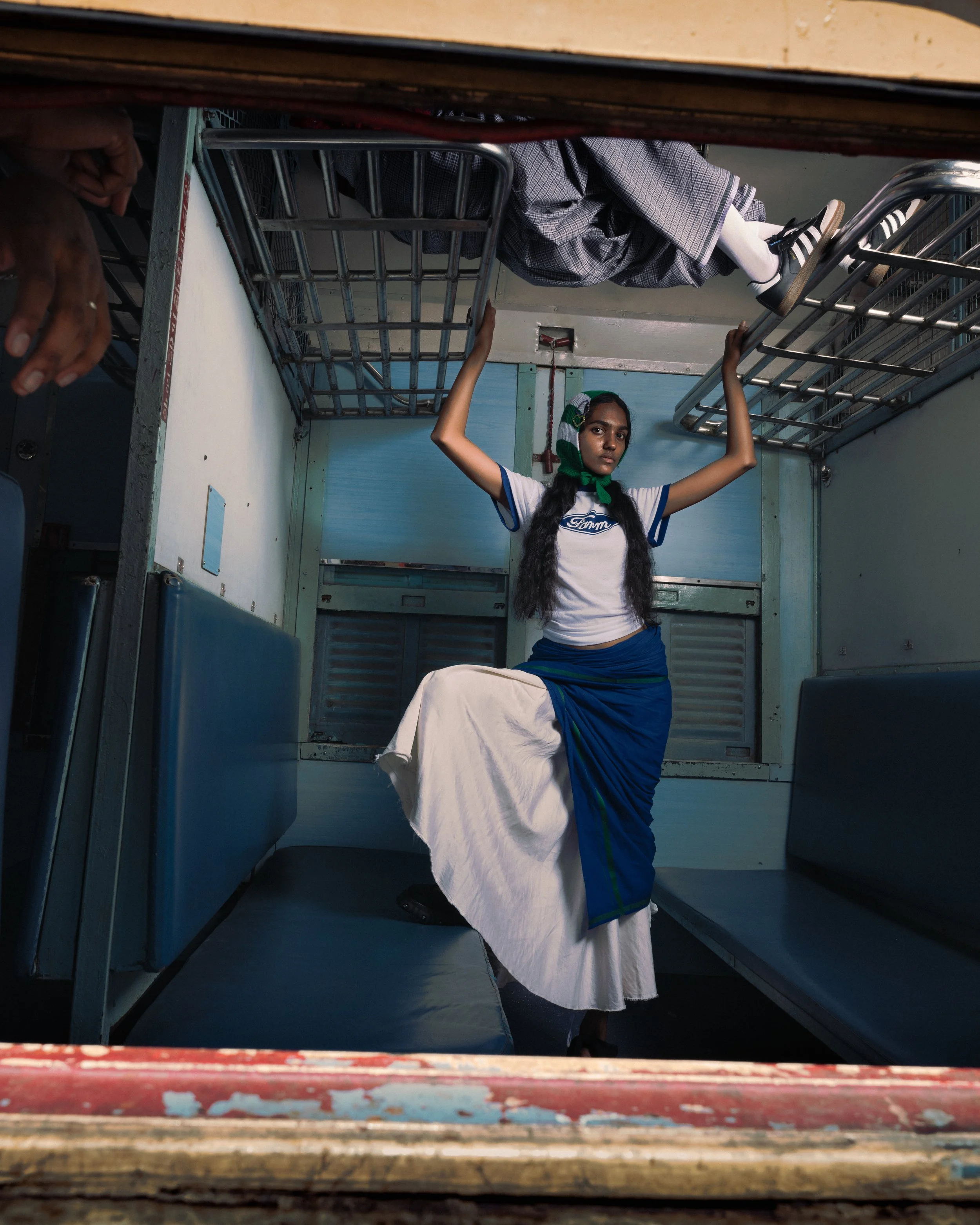 Une jeune femme portant une tenue blanche et bleue, avec une écharpe verte autour du cou, se tient à l'intérieur d'un train, debout sur un siège avec des mains agrippant le plafond du véhicule.
