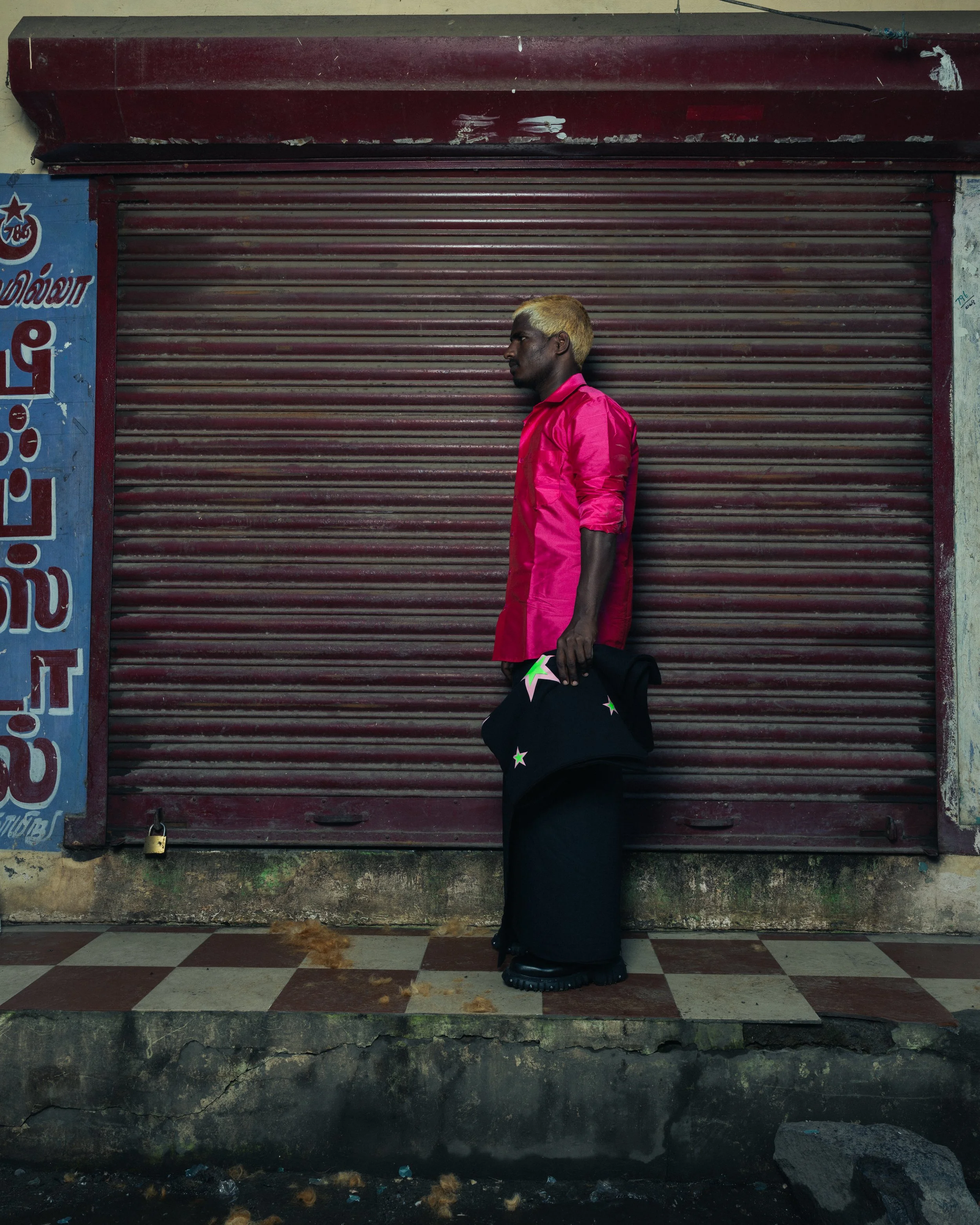 Jeune homme debout devant un volet roulant fermé, portant une chemise rose et un pantalon noir, tenant un vêtement avec des étoiles noires et vertes.