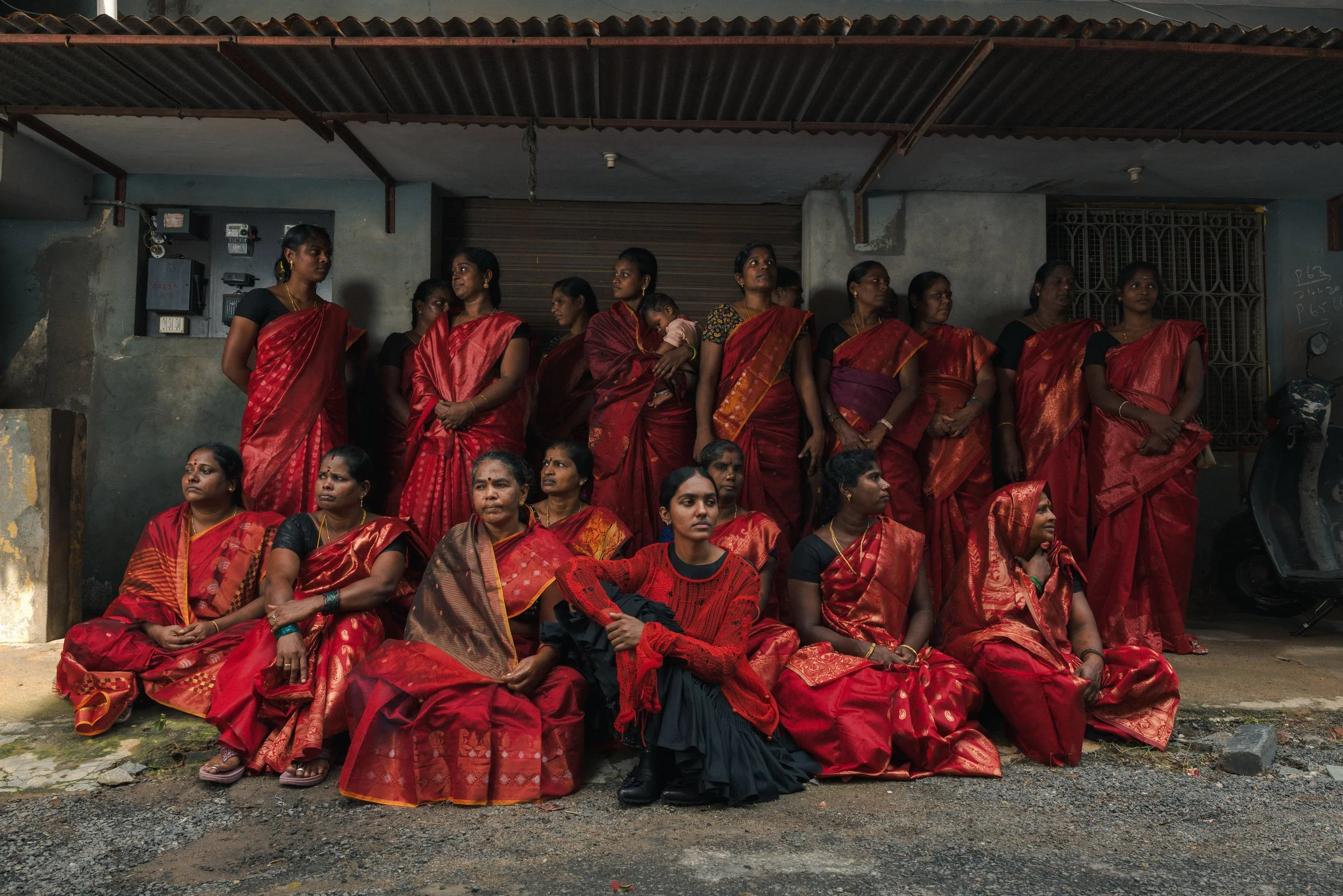 Groupe de femmes indiennes portant des saris rouges, certaines assises par terre et d'autres debout, devant un bâtiment.