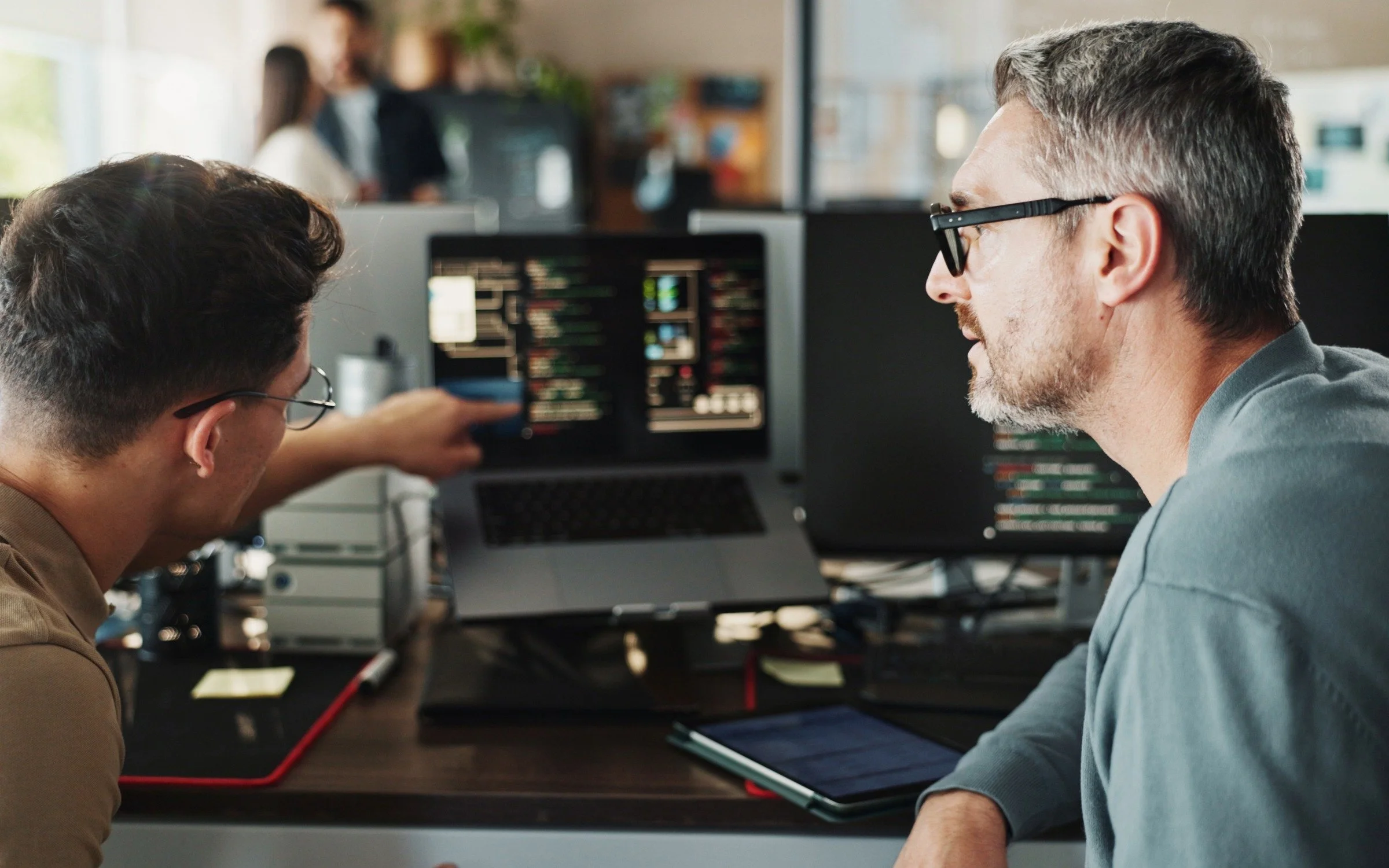 Two men with glasses working in a tech office, looking at computer screens displaying code, with one pointing at a monitor and a tablet on the desk.