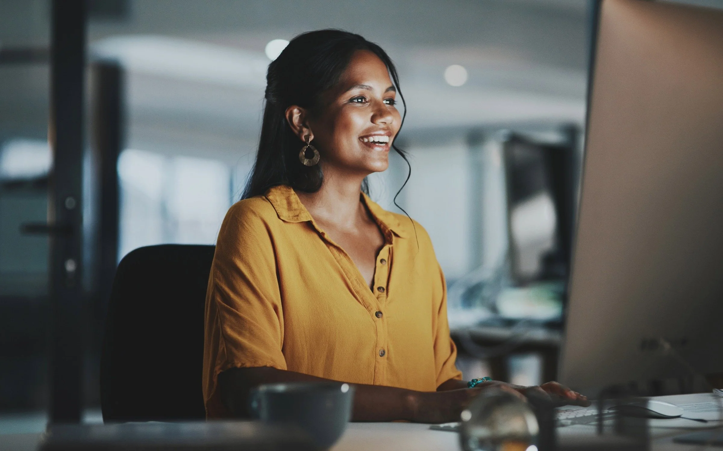 A woman wearing a mustard yellow blouse and large earrings is working at her desk using a computer in an office setting.