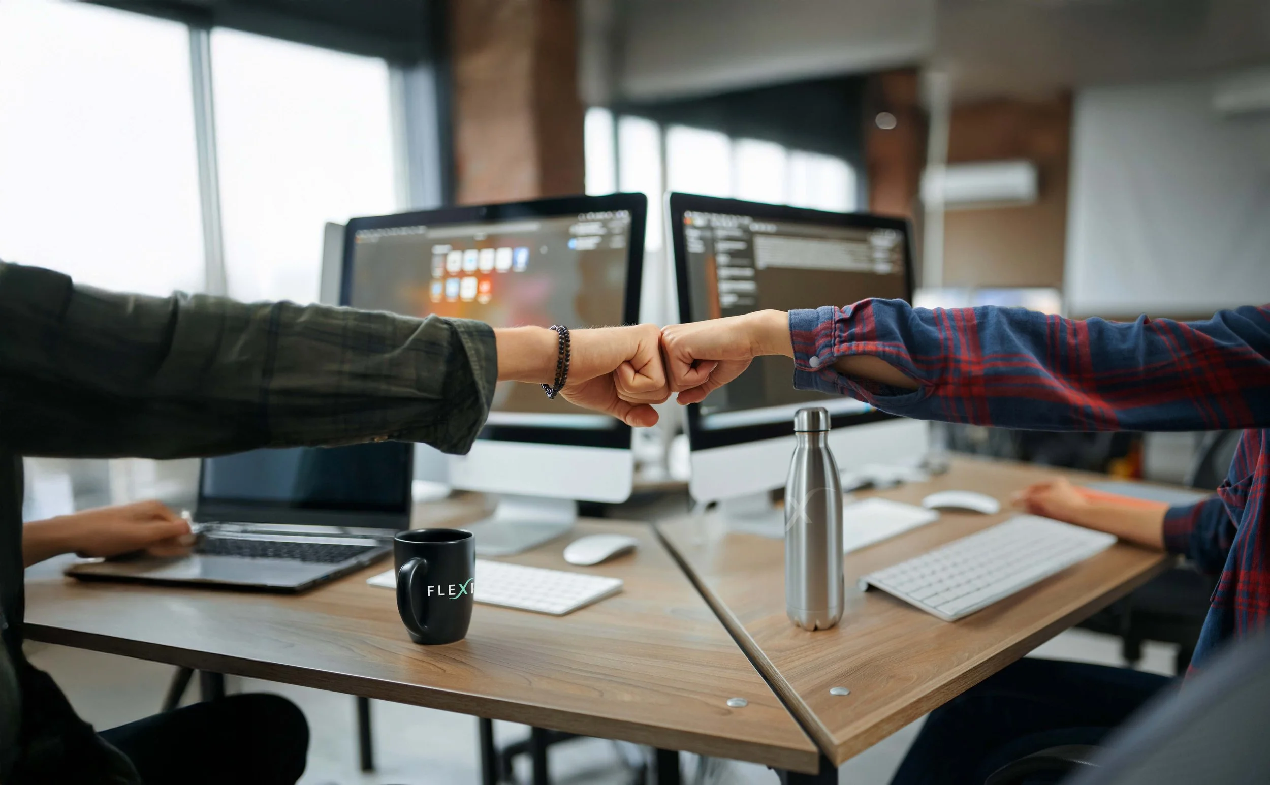 Two people giving each other fist bumps in an office with computers and monitors on the desk.