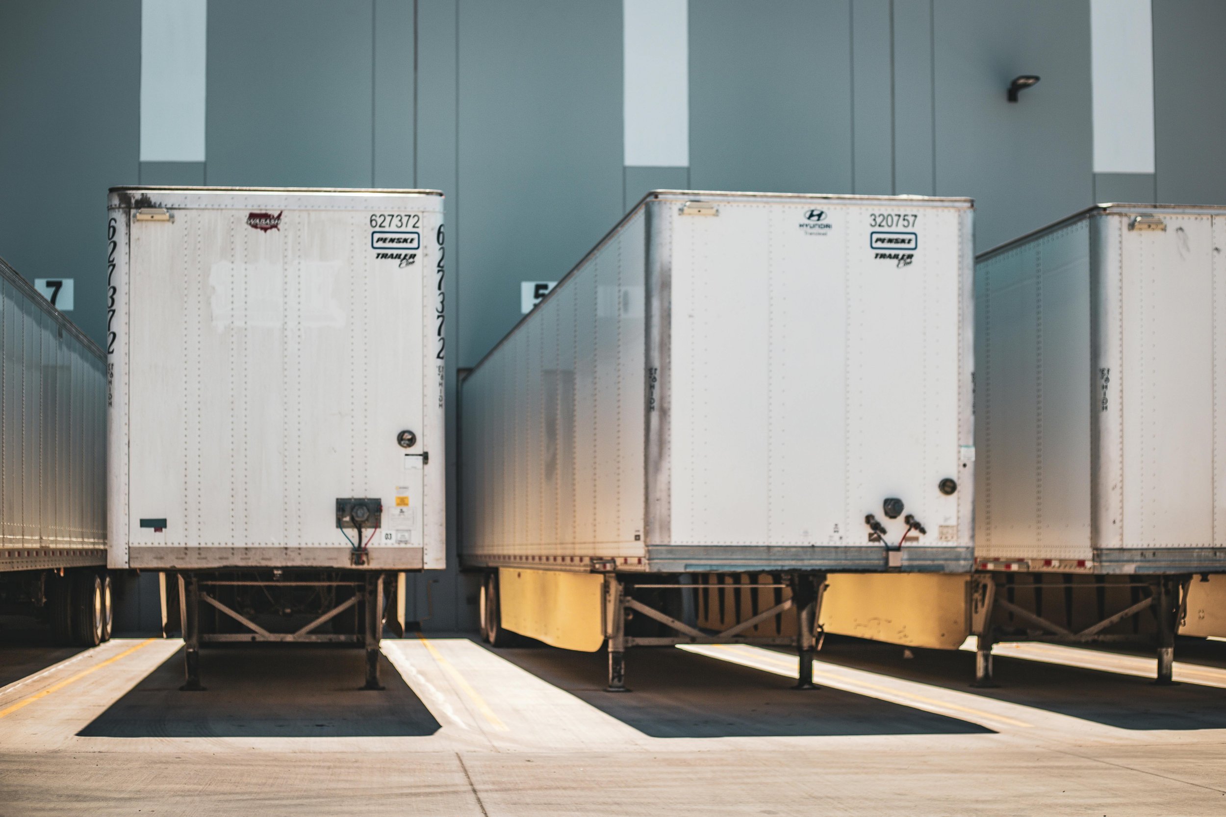 Three semi-trailers parked side by side in a lot, with a large industrial building in the background.