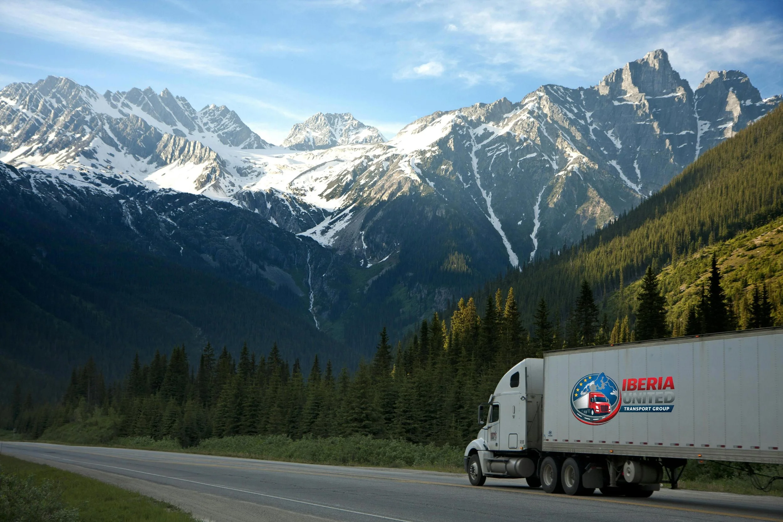 A white semi-truck with Iberia United Transport Group logo driving on a highway surrounded by dense green pine trees, with snow-capped mountain peaks and a partly cloudy sky in the background.